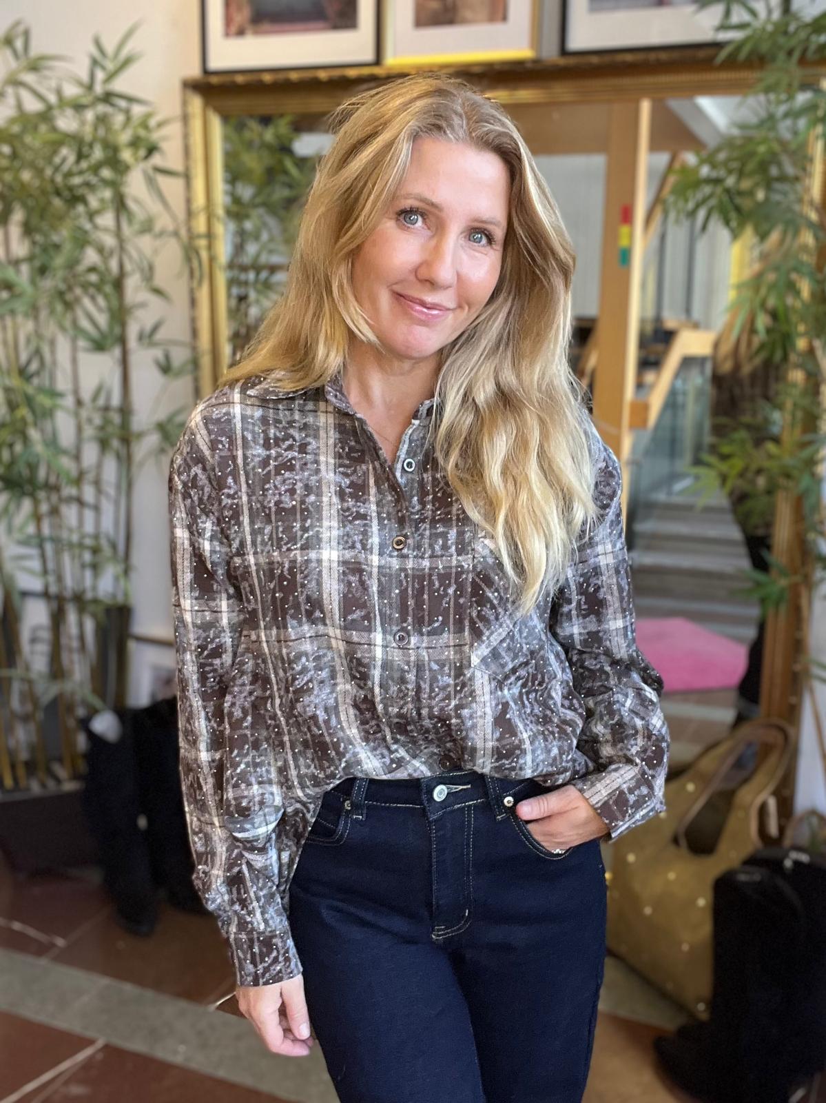 Leende kvinna med långt blont hår står inomhus och poserar med ena handen i fickan. Hon bär en mönstrad, rutig skjorta och mörka jeans; bakom syns spegel, växter och väskor i en hemlik miljö.