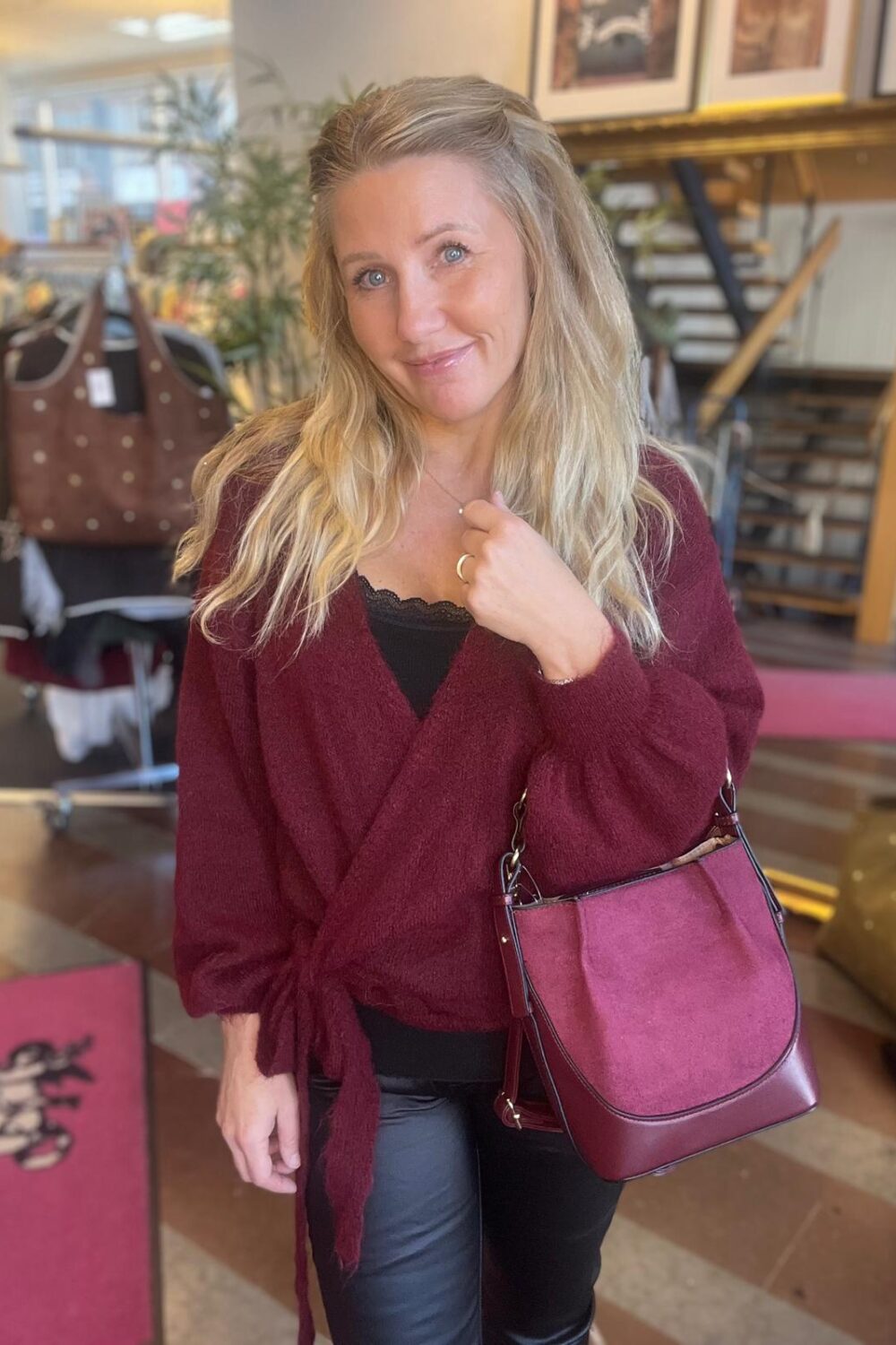 Leende kvinna med långt blont hår poserar inomhus och håller i ett halsband. Hon bär vinröd stickad omlottkofta och svarta byxor, med en vinröd handväska över armen. Bakgrunden visar butiksmiljö med kläder och trappa.