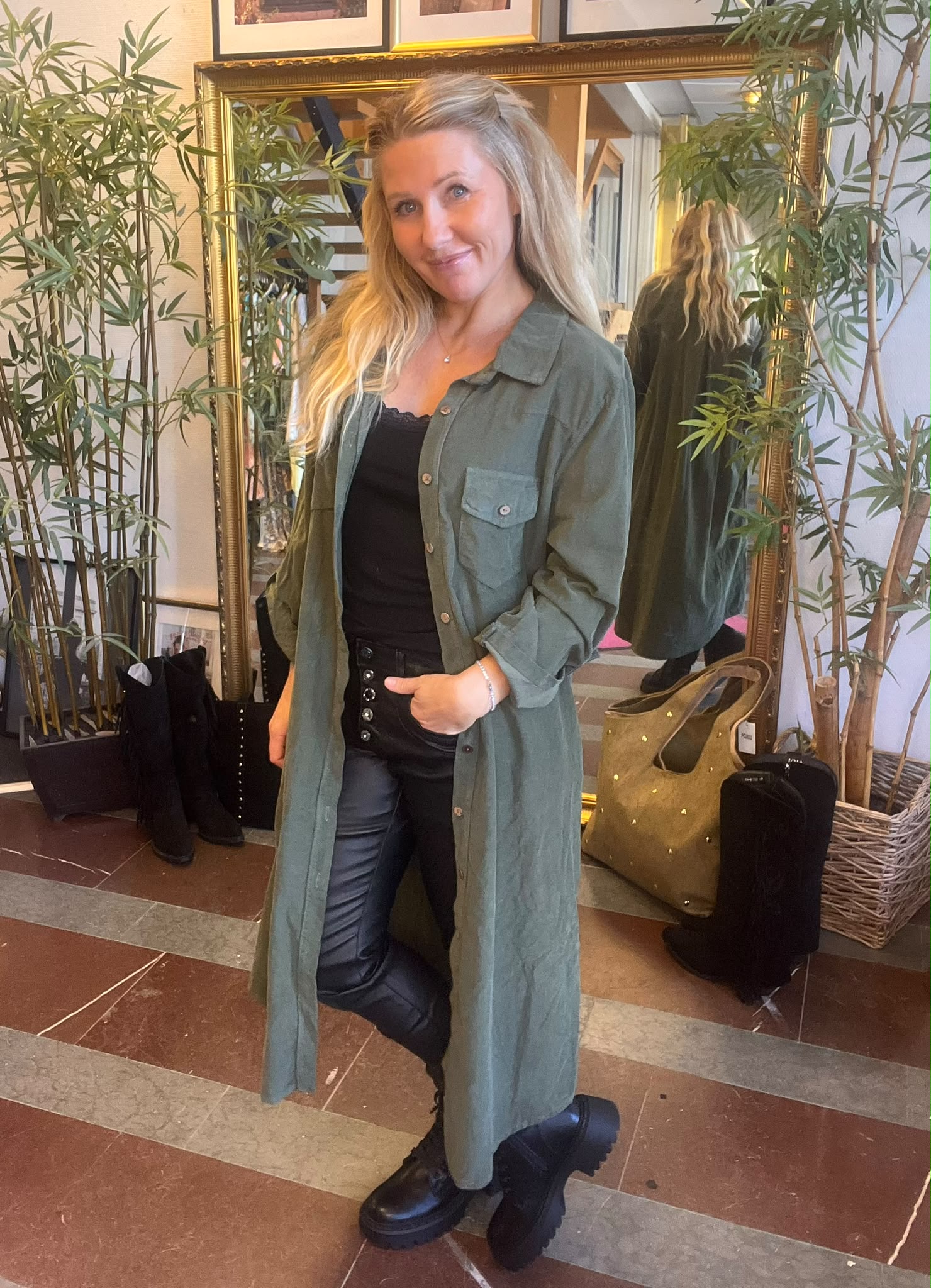 Kvinna med långt blont hår står och ler med ena handen i fickan, klädd i lång grön skjortklänning över svart topp, mörka byxor och grova svarta kängor. Bakom syns spegel, växter och väskor.