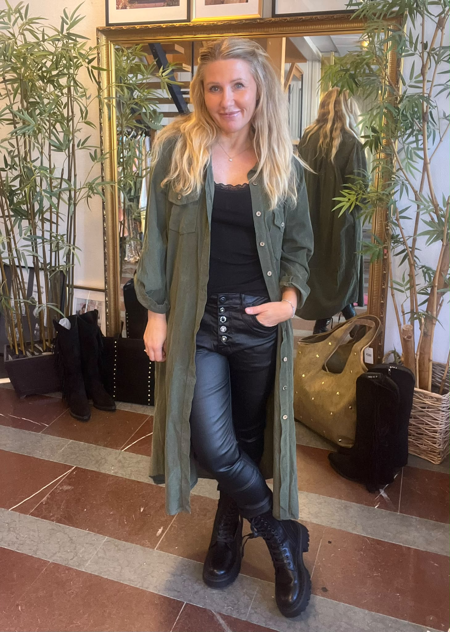 Leende kvinna poserar med handen i fickan framför en stor guldramsspegel, omgiven av höga gröna växter. Hon bär lång grön skjortklänning, svart topp och läderbyxor med knappar samt svarta kängor i butiksmiljö.