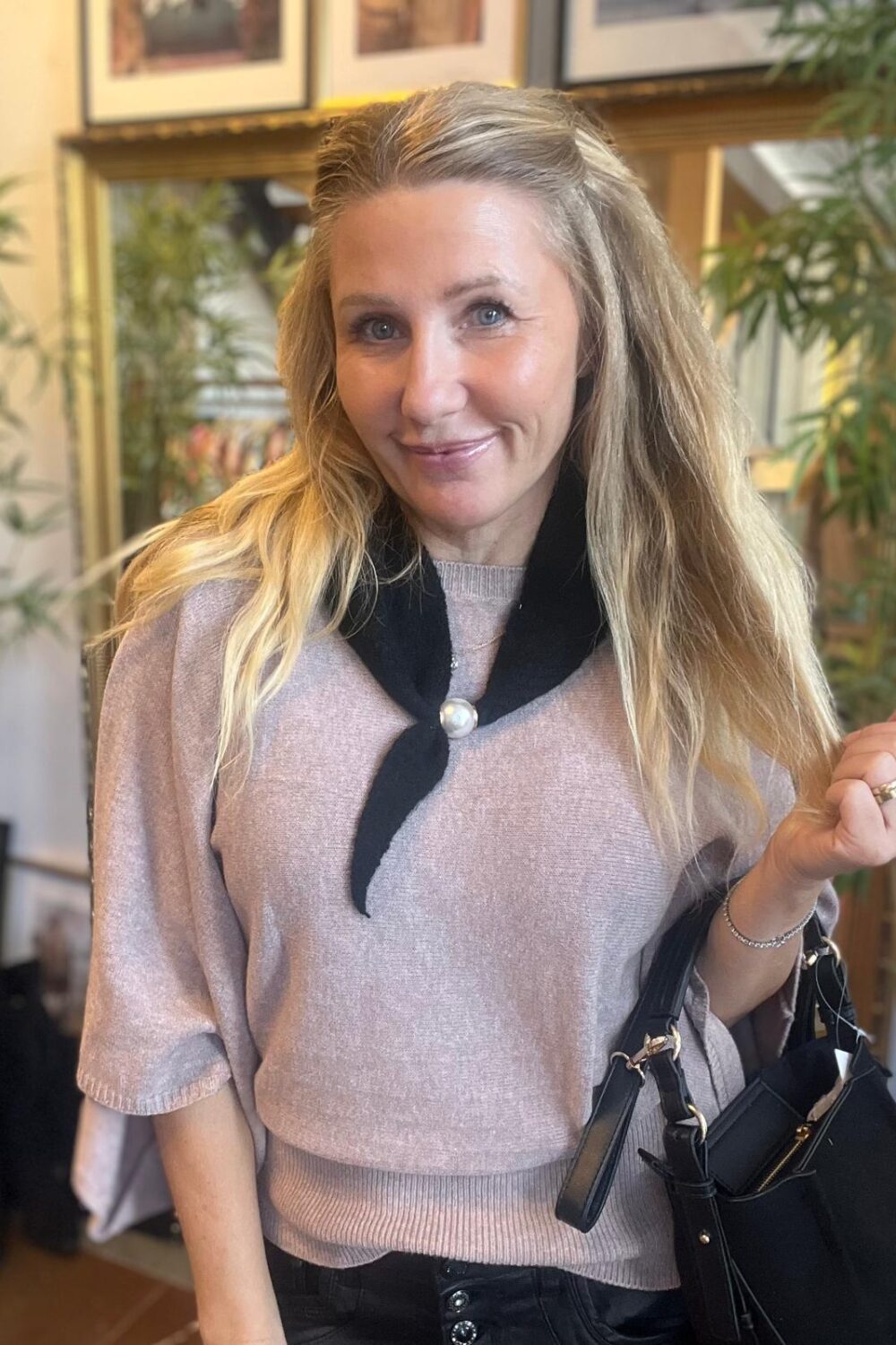 Leende blond kvinna tittar in i kameran och håller en svart handväska i handen. Hon bär ljusrosa stickad tröja och svart halsduk med pärlknapp, inomhus framför växter och inramade tavlor.