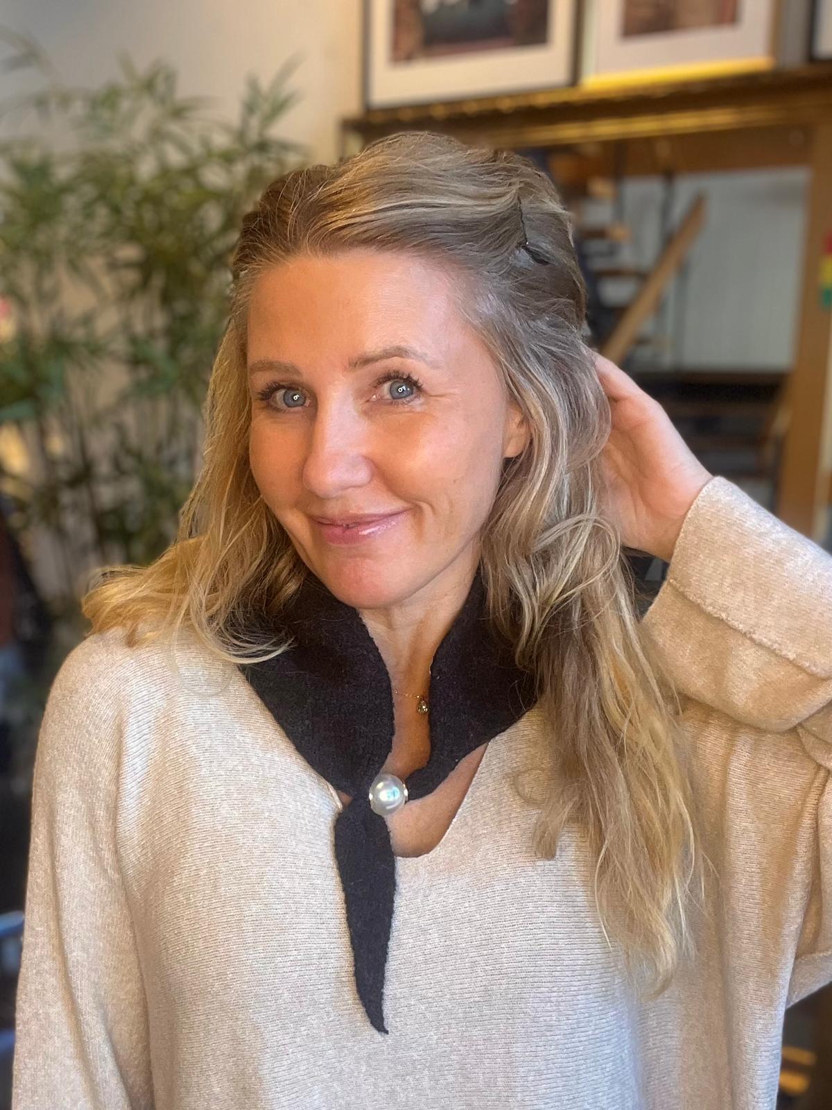 Leende kvinna med långt blont hår tittar mot kameran och lyfter ena handen bakom huvudet. Hon bär en ljus tröja med mörk krage och pärlknapp, i ett hemtrevligt rum med växt och tavlor.