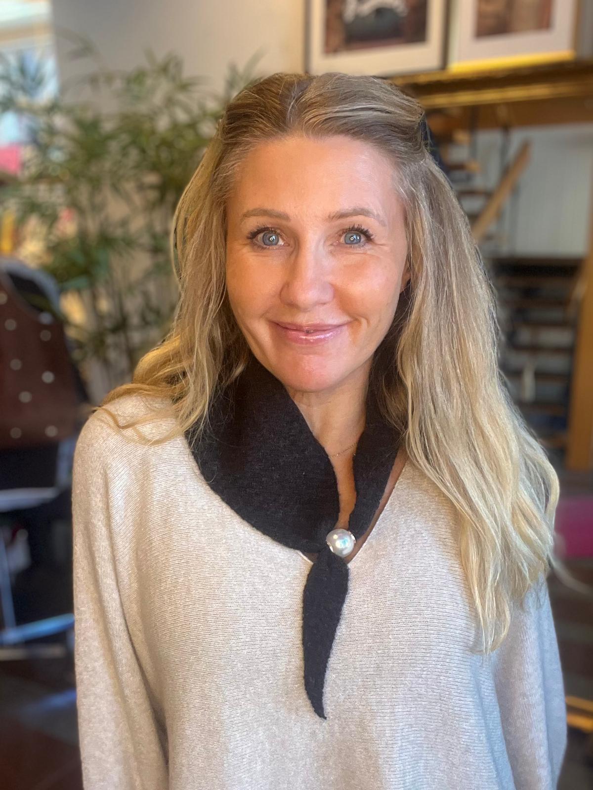 Leende kvinna med långt blont hår tittar in i kameran, klädd i ljus tröja med mörk krage och pärlknapp. Hon står inomhus i ett rum med växt och möbler i mjuk bakgrundsoskärpa.