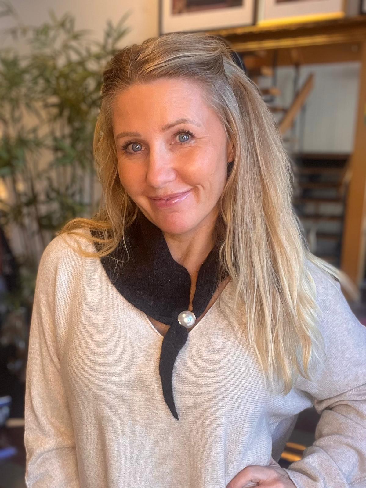 Leende kvinna med långt blont hår tittar in i kameran och lutar sig lätt framåt. Hon bär ljus tröja och mörk sjal med pärlknapp, inomhus i en varm miljö med växt och trappa i bakgrunden.