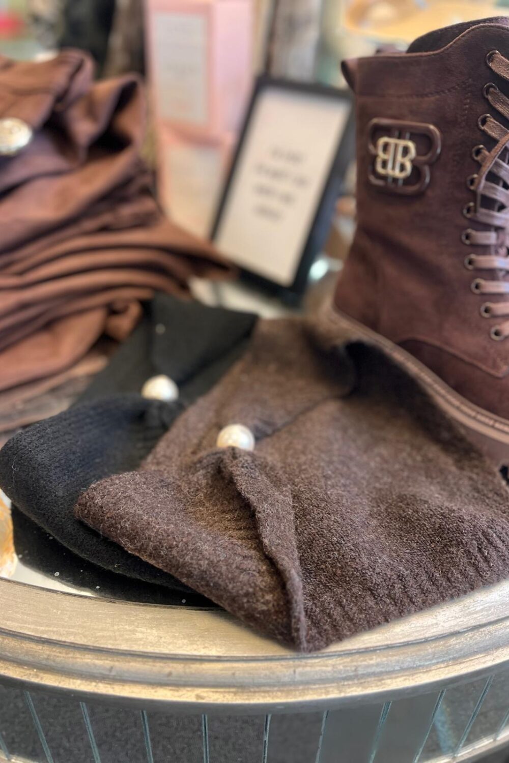 Brun snörkänga med dekorativt BB-emblem står uppställd i ett butikskyltfönster. Framför ligger en mörk ullmössa med pärldetaljer och ett svart plagg, medan hopvikta bruna textilier och suddiga skyltar syns i bakgrunden.