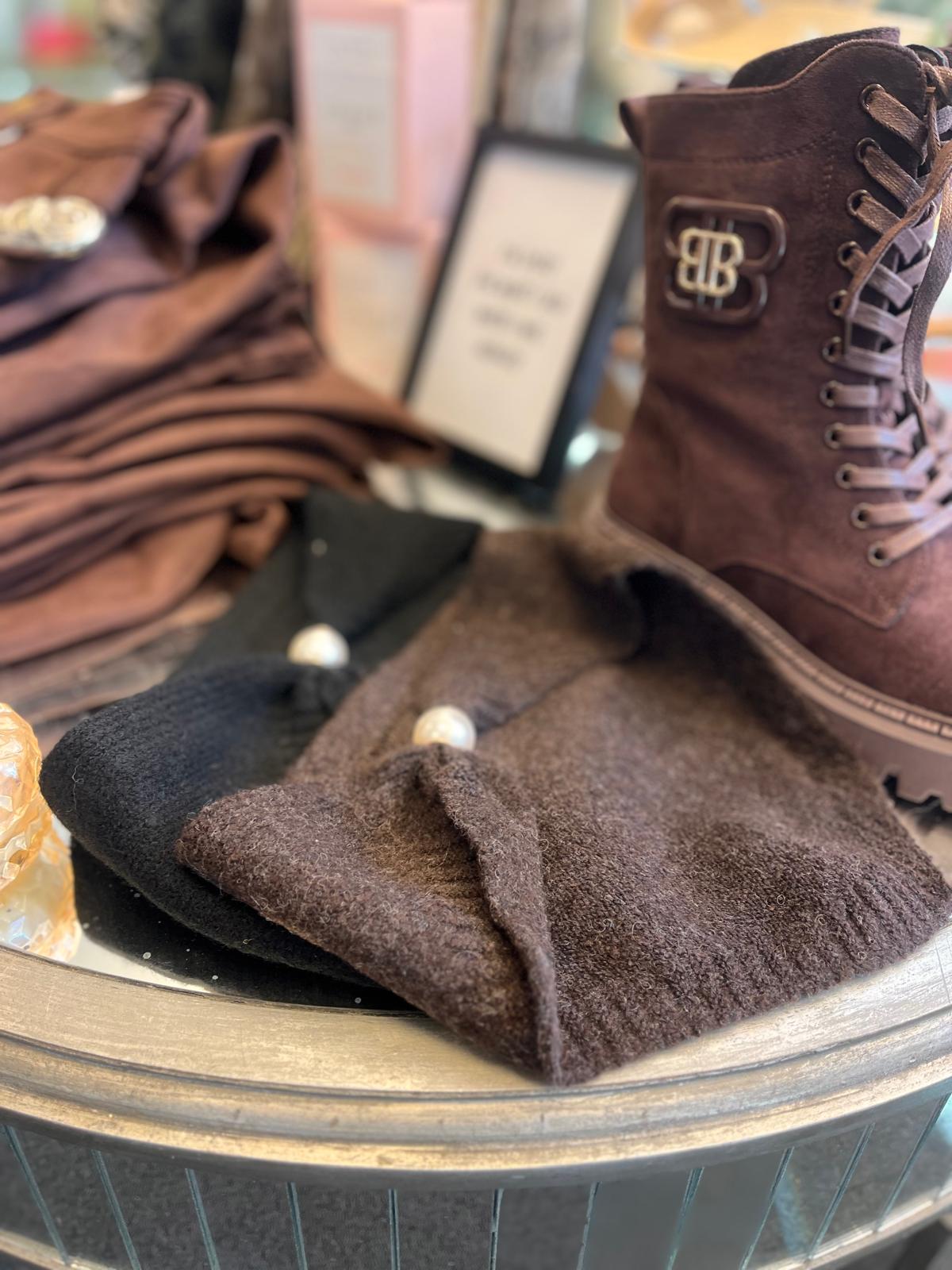 Brun snörkänga med dekorativt BB-emblem står uppställd i ett butikskyltfönster. Framför ligger en mörk ullmössa med pärldetaljer och ett svart plagg, medan hopvikta bruna textilier och suddiga skyltar syns i bakgrunden.