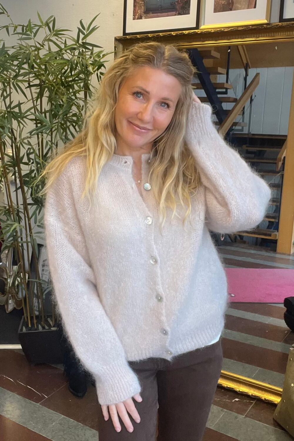 Leende kvinna med långt blont hår poserar och lyfter ena handen mot håret. Hon bär en ljus, stickad kofta med knappar och bruna byxor, i en inomhusmiljö med stor guldramsspegel och växt i bakgrunden.