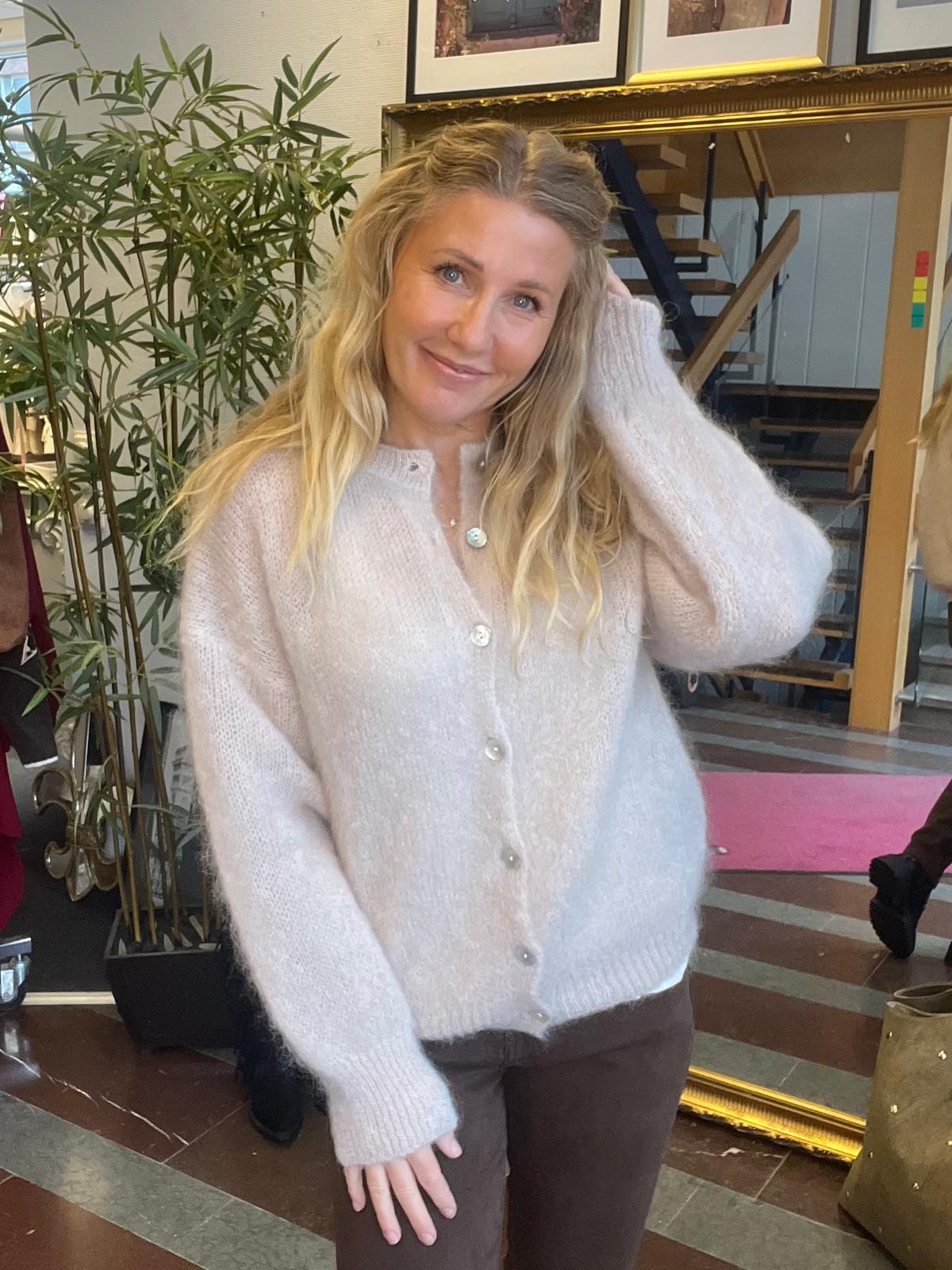 Leende kvinna med långt blont hår poserar och lyfter ena handen mot håret. Hon bär en ljus, stickad kofta med knappar och bruna byxor, i en inomhusmiljö med stor guldramsspegel och växt i bakgrunden.