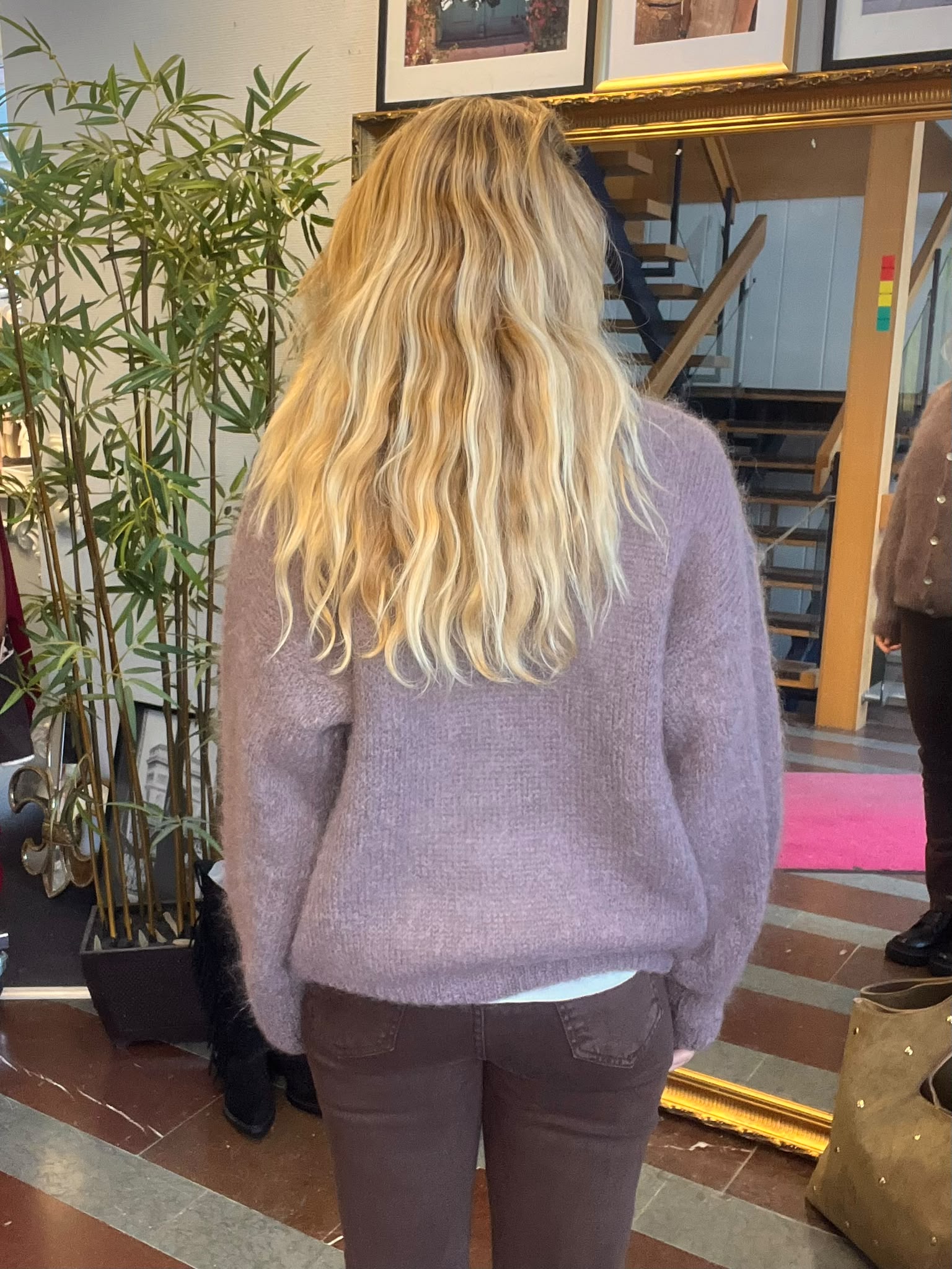 Person med långt, vågigt blont hår står med ryggen mot kameran i en ljuslila stickad tröja och mörka byxor, inomhus vid en stor guldspegel, bambuväxt och en trappa; en annan person syns delvis till höger.