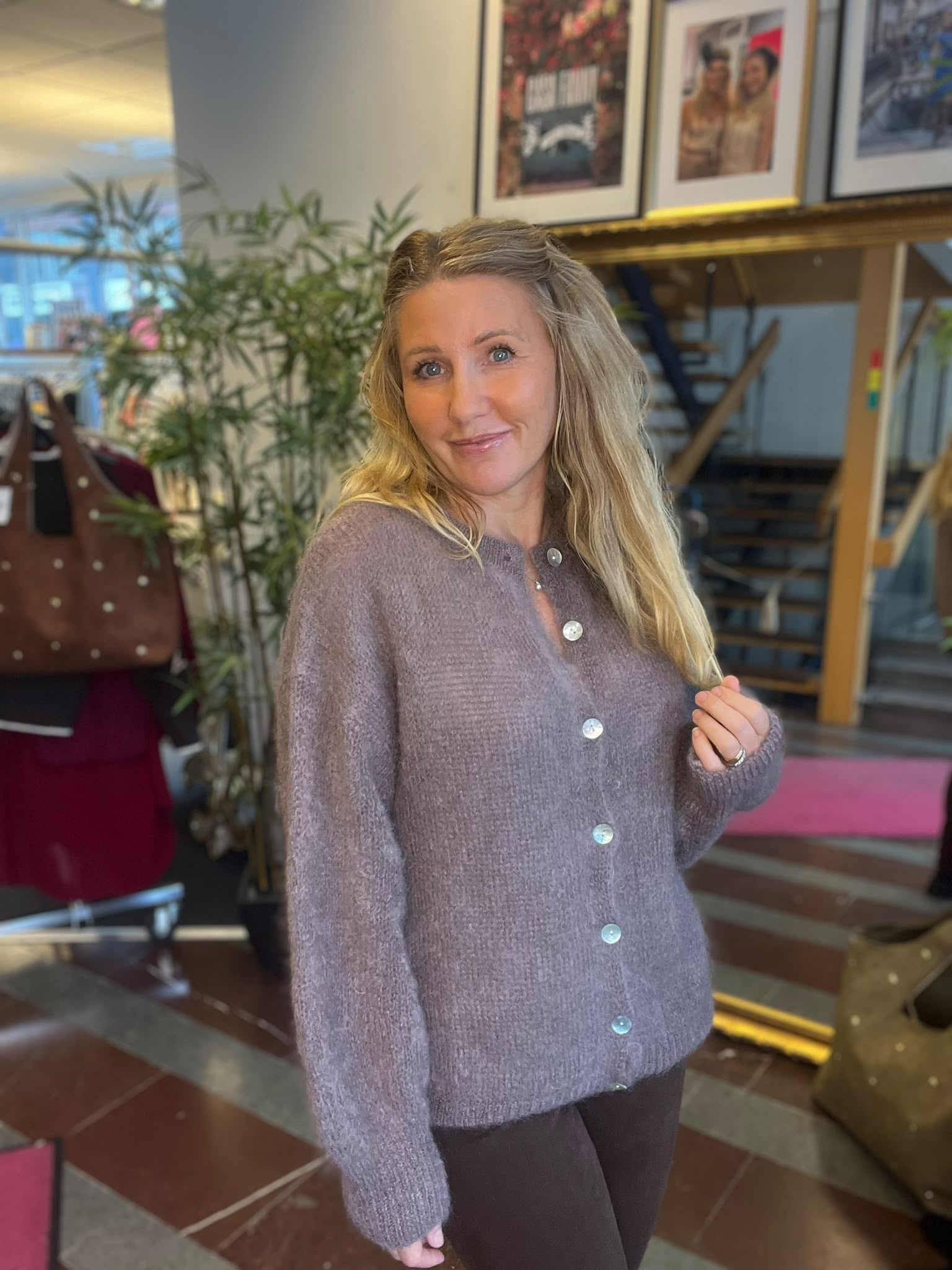 Blond kvinna i lila stickad kofta står och ler mot kameran, med ena handen vid håret. Inomhusmiljö med stor grön växt, trappa och inramade affischer på väggen i bakgrunden.