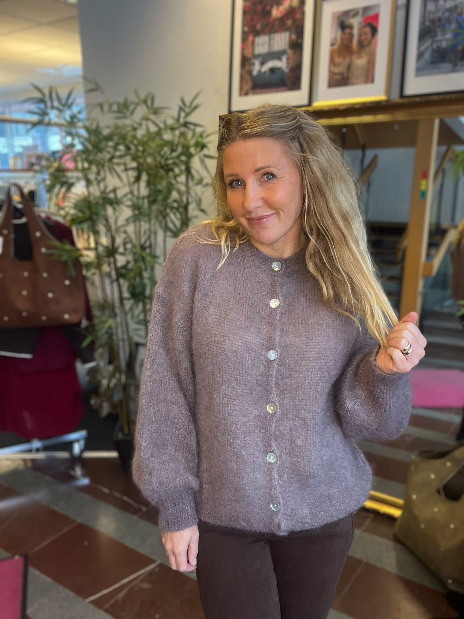 Leende kvinna med långt blont hår poserar inomhus och håller upp handen som om hon visar en ring. Hon bär en lila stickad kofta. I bakgrunden syns en stor växt, stolar och inramade foton på väggen.