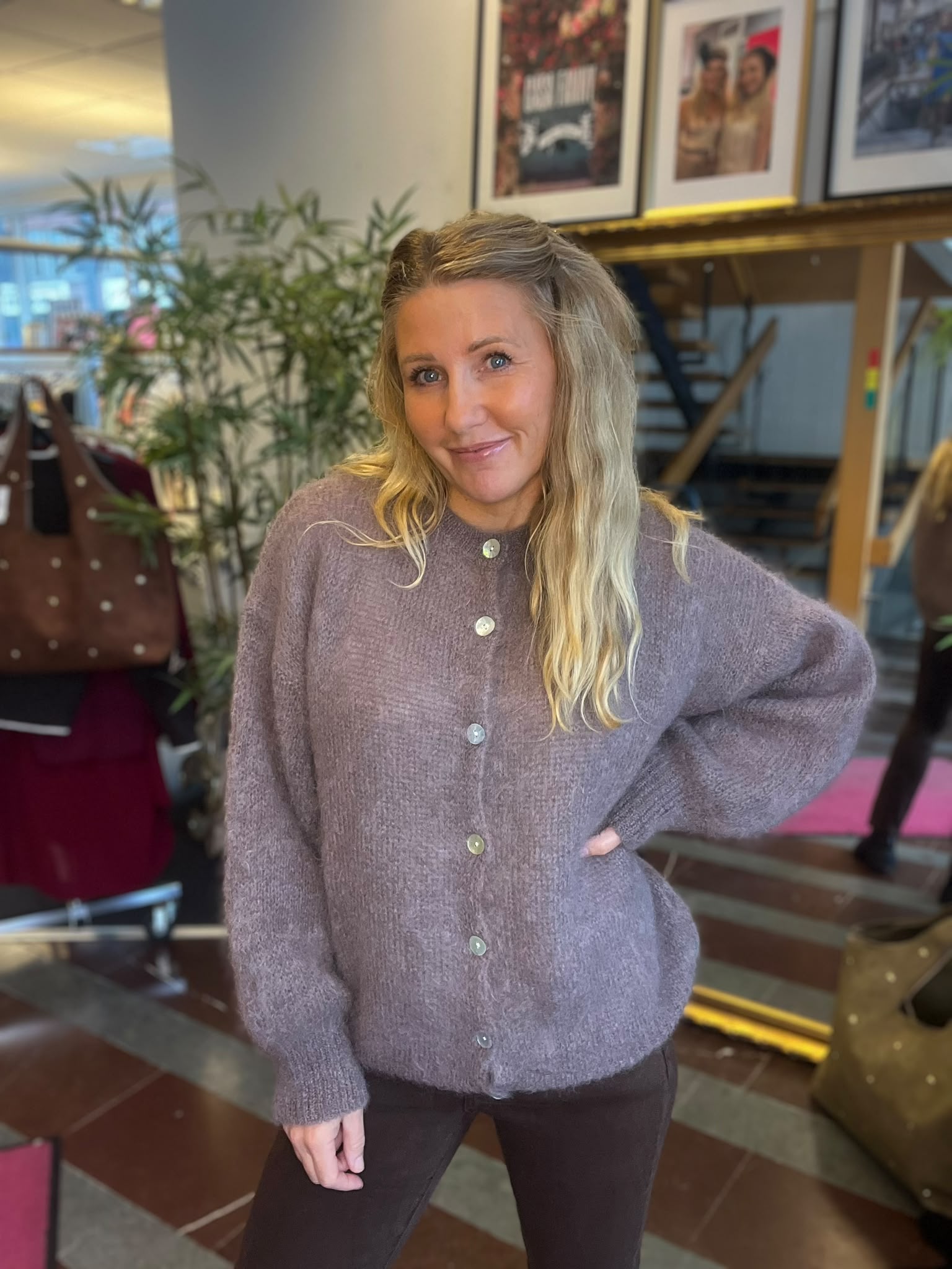Leende kvinna med långt blont hår står med handen i sidan och tittar mot kameran. Hon bär en lila stickad kofta med knappar, i en inomhusmiljö med växter, trappa och inramade foton i bakgrunden.