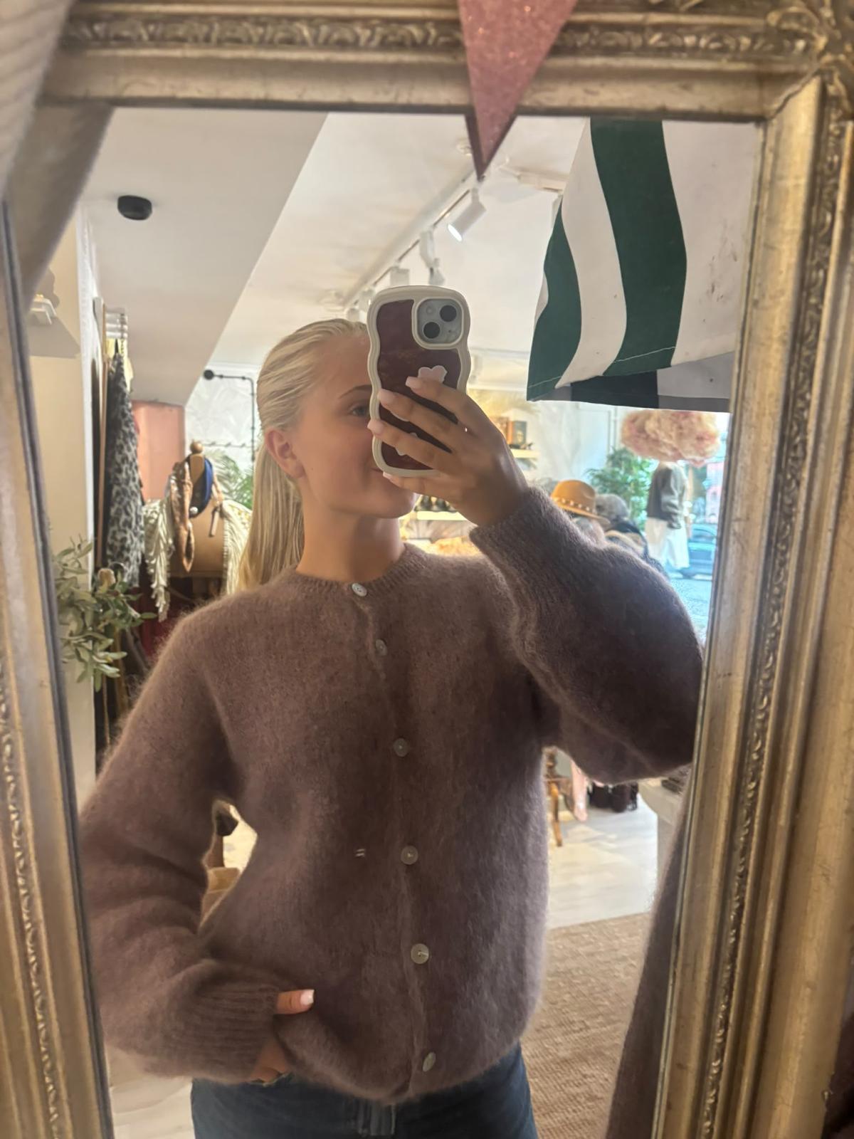 Person tar en spegelselfie i en butik, håller mobilen framför ansiktet och bär en mjuk, brun stickad kofta med knappar och jeans. I bakgrunden syns klädställningar, hattar och inredning i butiken.