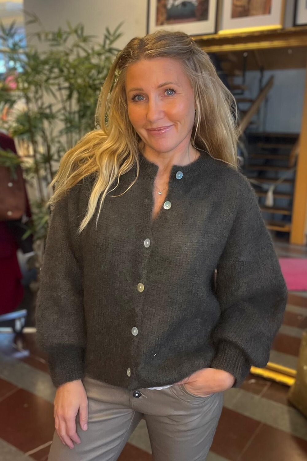 Leende kvinna med långt blont hår poserar inomhus med ena handen i fickan. Hon bär mörkgrå knappad kofta och ljusbruna byxor, med växter, röd stol och en trappa i bakgrunden.