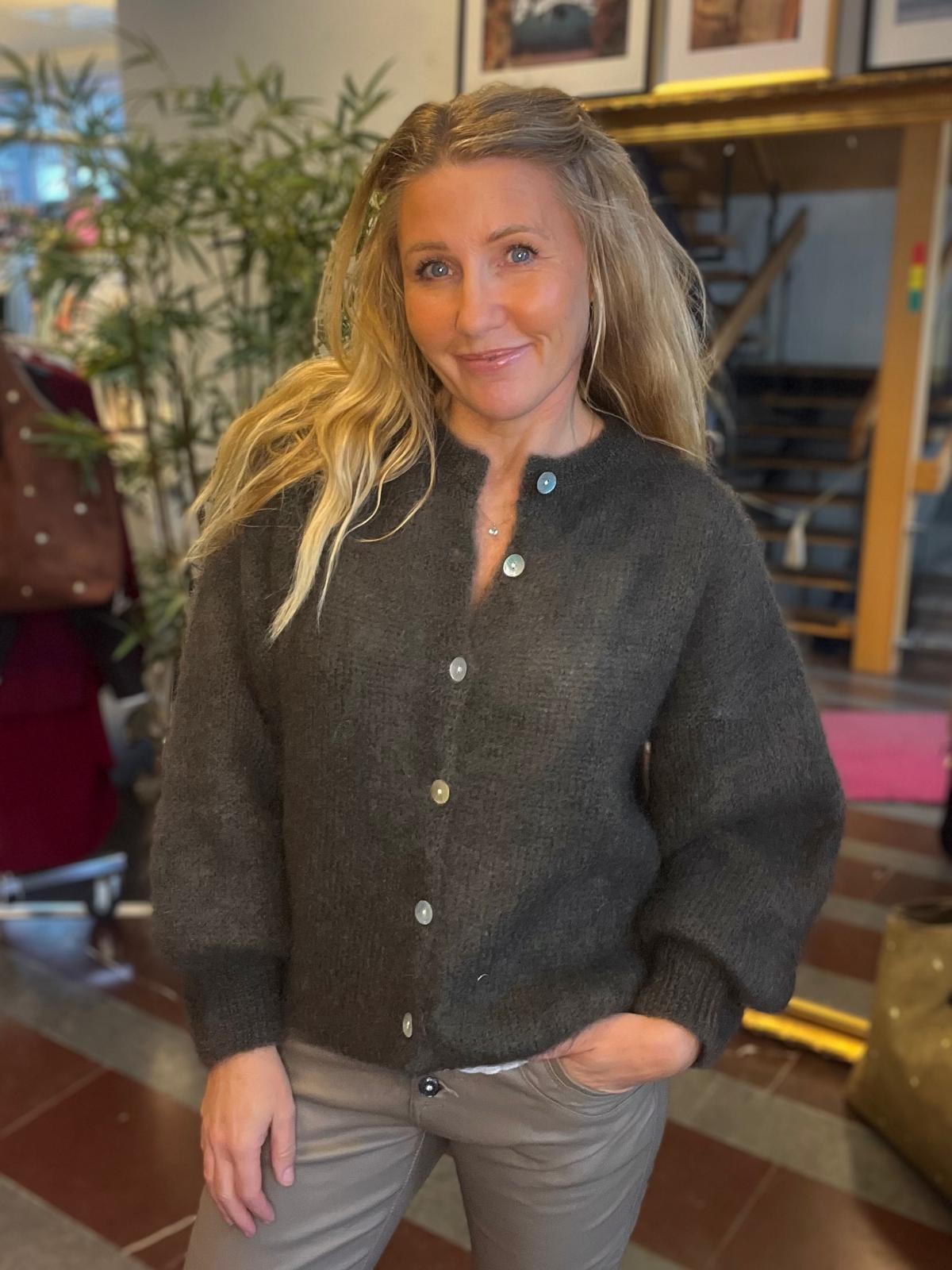 Leende kvinna med långt blont hår poserar inomhus med ena handen i fickan. Hon bär mörkgrå knappad kofta och ljusbruna byxor, med växter, röd stol och en trappa i bakgrunden.