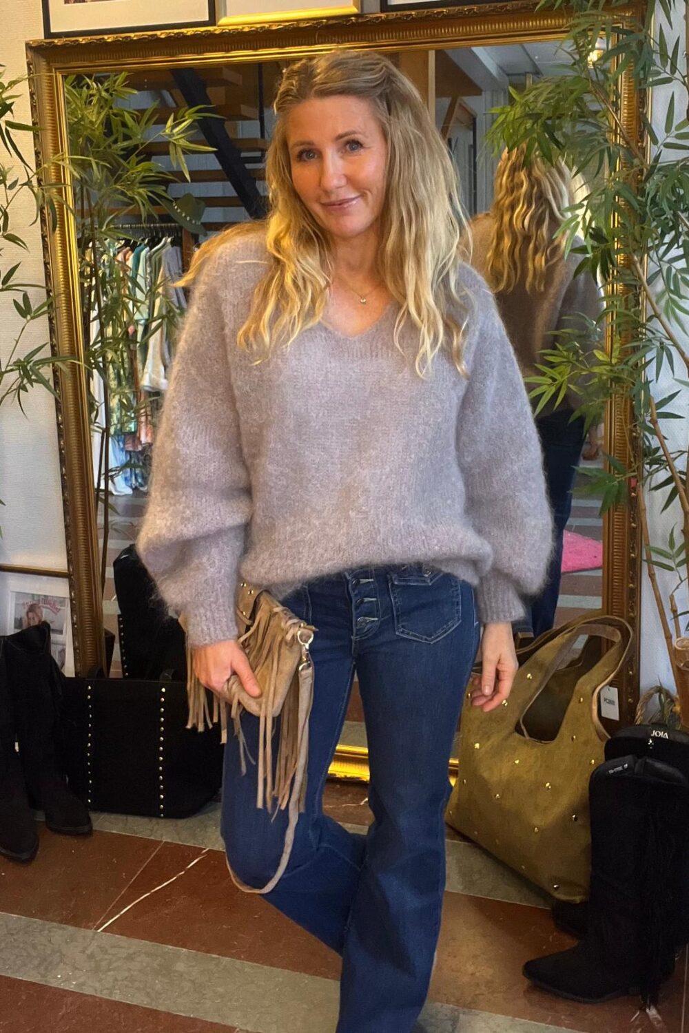 Kvinna med långt blont hår poserar och ler framför en stor guldramad spegel, klädd i lila fluffig tröja och blå jeans, med fransad handväska i handen. Bakom syns klädställning, stövlar, stora väskor och gröna växter.