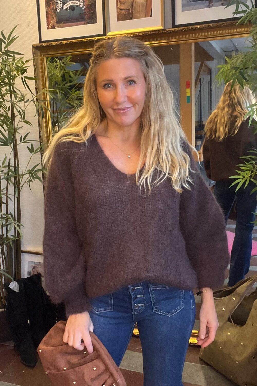 Kvinna med långt blont hår står och ler mot kameran, iklädd lila stickad tröja och blå jeans med knappar. Hon håller en brun väska i en inomhusmiljö med spegel, växter och tavlor.