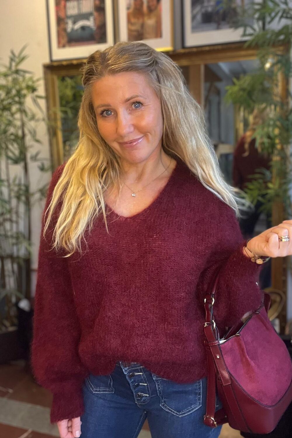 Leende kvinna med långt blont hår poserar inomhus och håller upp ena handen, med en vinröd handväska över armen. Hon bär vinröd stickad tröja och jeans; bakom syns spegel, växter och inramade bilder.