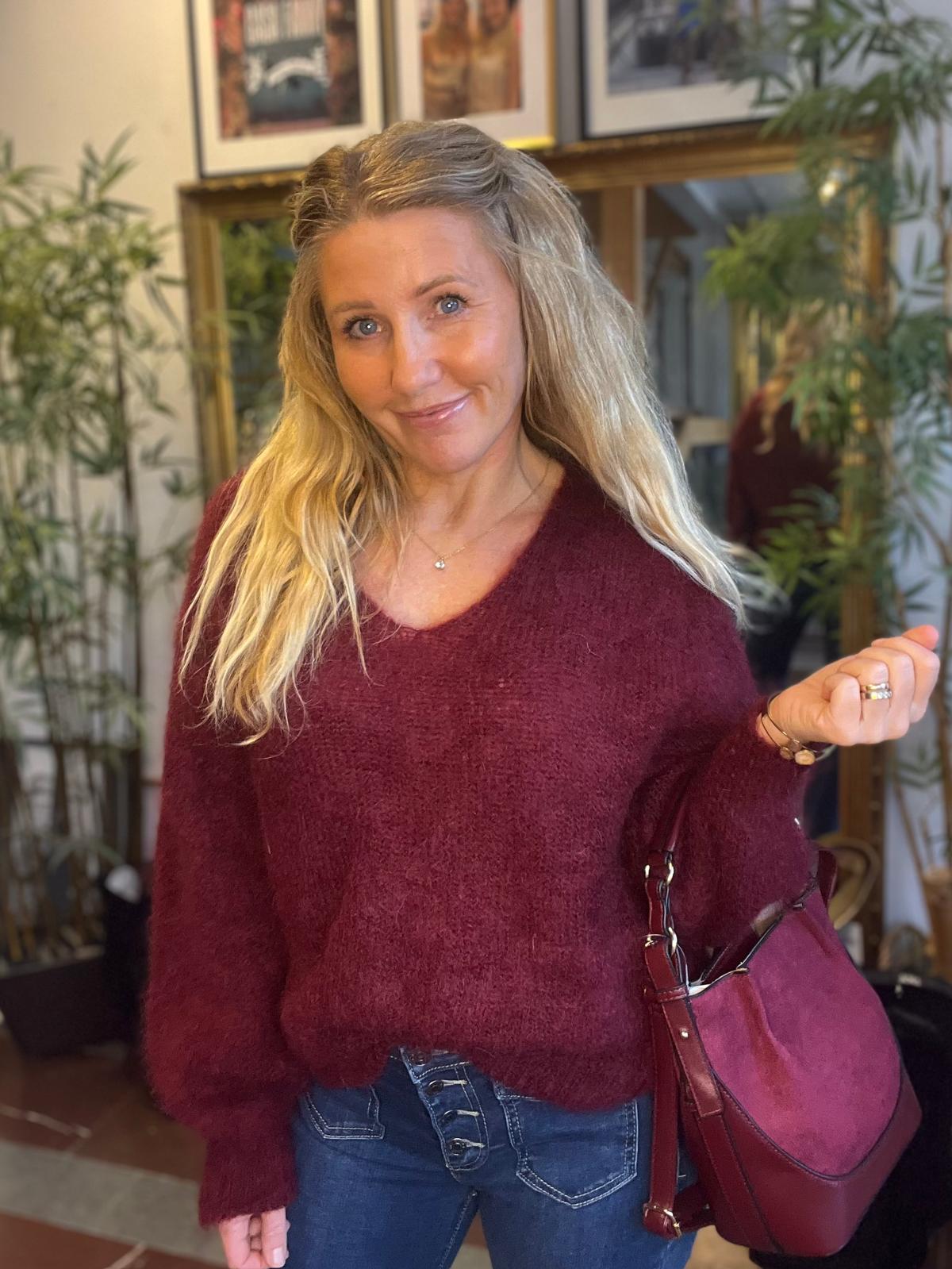 Leende kvinna med långt blont hår poserar inomhus och håller upp ena handen, med en vinröd handväska över armen. Hon bär vinröd stickad tröja och jeans; bakom syns spegel, växter och inramade bilder.