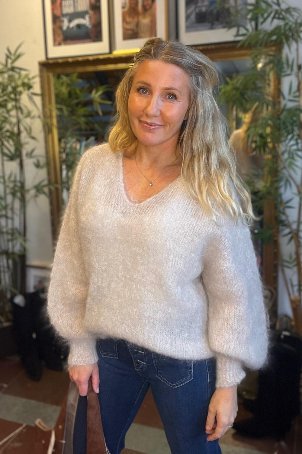 Kvinna med långt blont hår står och ler mot kameran, klädd i ljus stickad tröja och blå jeans. Hon håller en brun väska i handen, i ett rum med gröna växter och inramade tavlor.