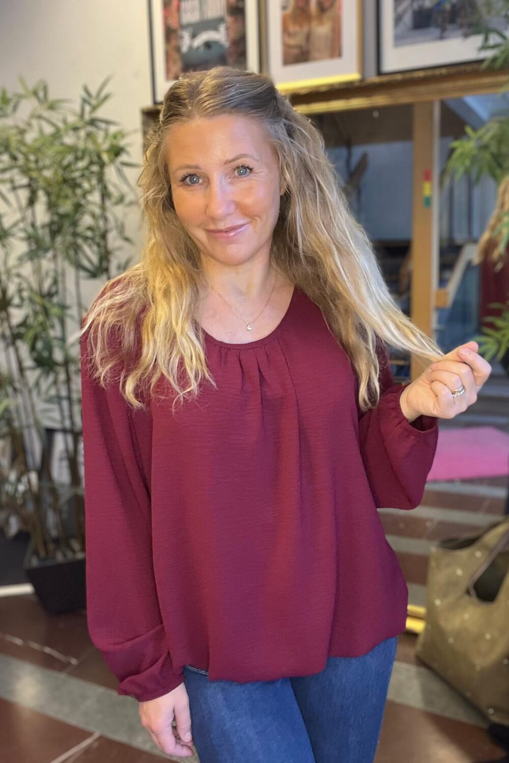 Leende kvinna med långt blont hår står inomhus och håller en hårslinga i handen. Hon bär vinröd blus och jeans; i bakgrunden syns gröna krukväxter, inramade bilder och en stor väska på golvet.