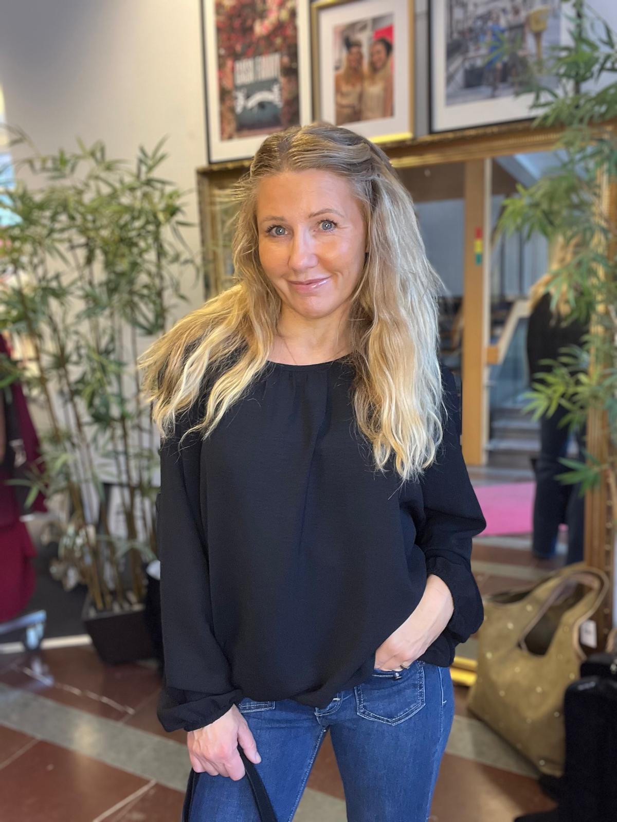 Leende kvinna med långt blont hår står inomhus och ser in i kameran, med ena handen i jeansfickan och den andra som håller en jacka. I bakgrunden syns krukväxter, inramade foton och en spegel.