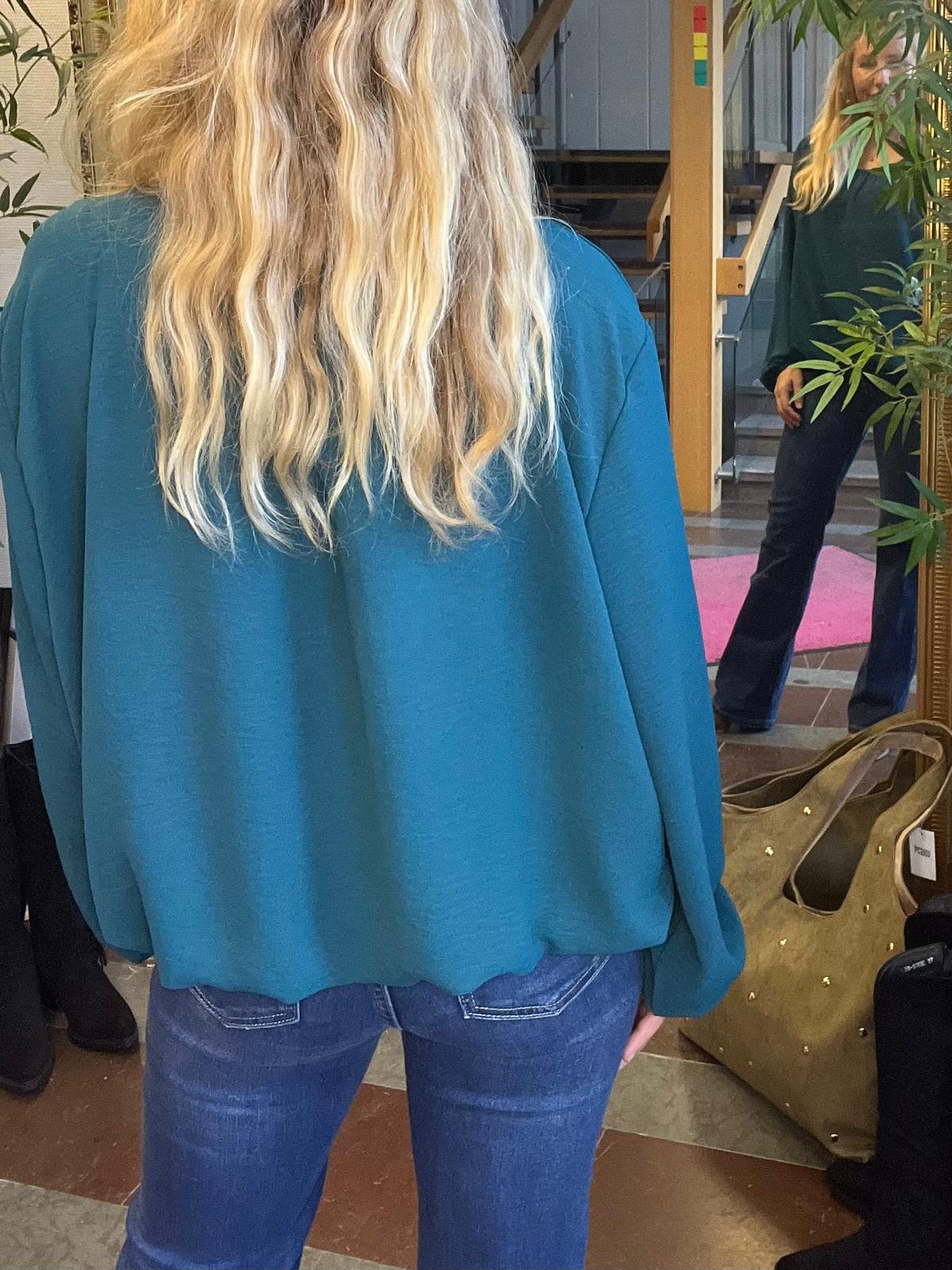 Kvinna med långt blont hår står med ryggen mot kameran och bär en turkos topp och jeans. Hon verkar posera framför en spegel; i spegeln syns hennes framsida i en hall med trappa och växter.