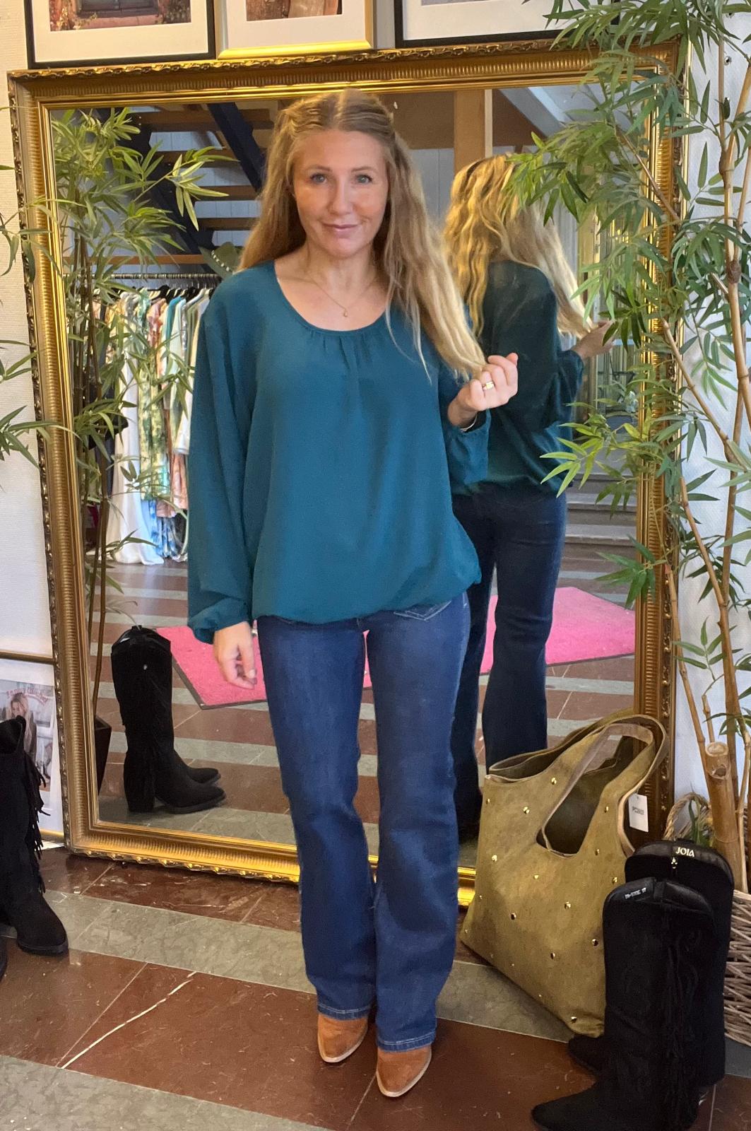 Kvinna står och poserar framför en stor guldramsspegel, klädd i petrolblå blus, blå jeans och bruna boots. Spegeln visar hennes rygg. Runt henne syns klädställning, svarta stövlar, stor väska och gröna växter.