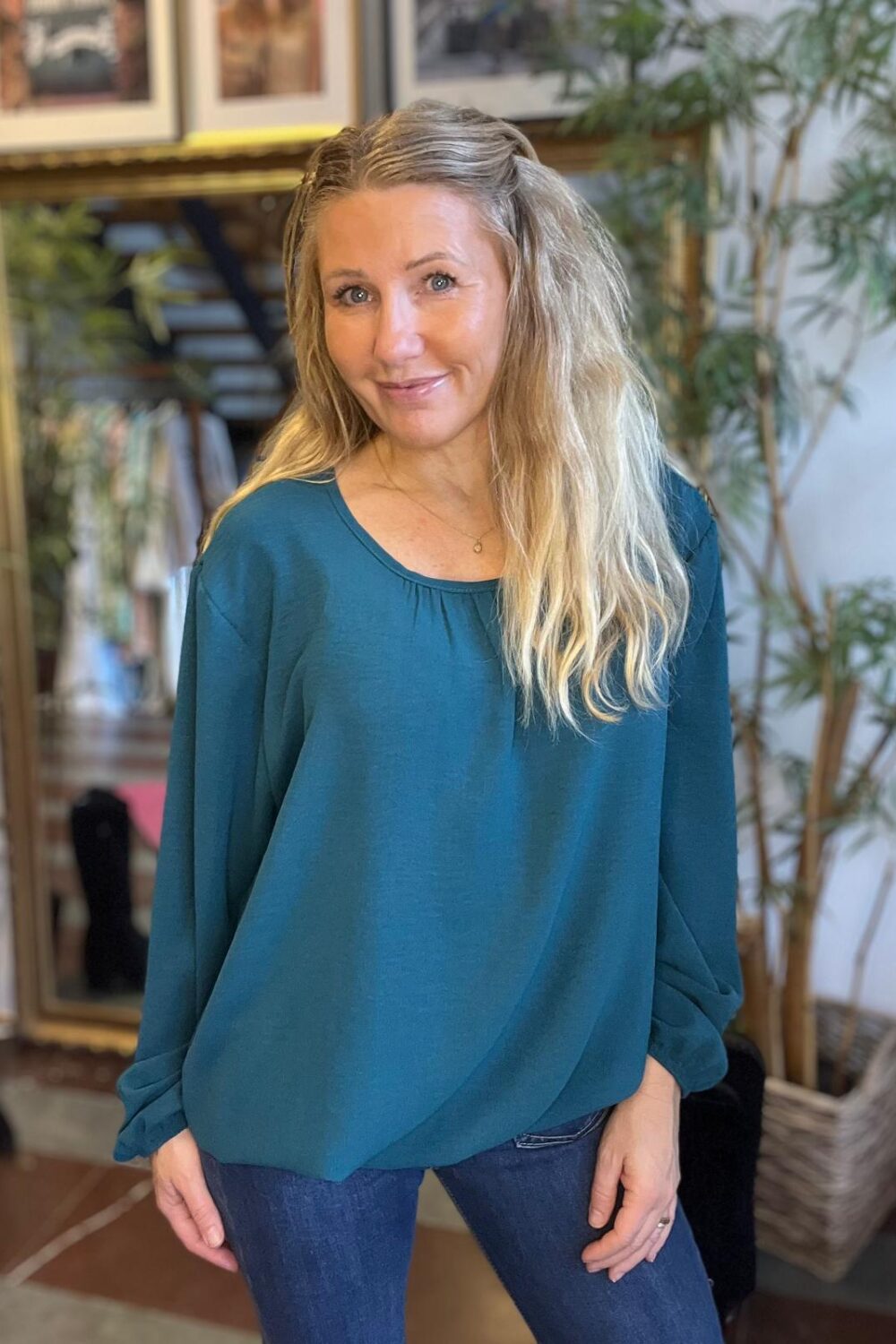 Kvinna med långt blont hår står och ler mot kameran, klädd i en petrolfärgad blus med långa ärmar och blå jeans. Hon poserar inomhus framför en stor spegel, växter och ramar på väggen.