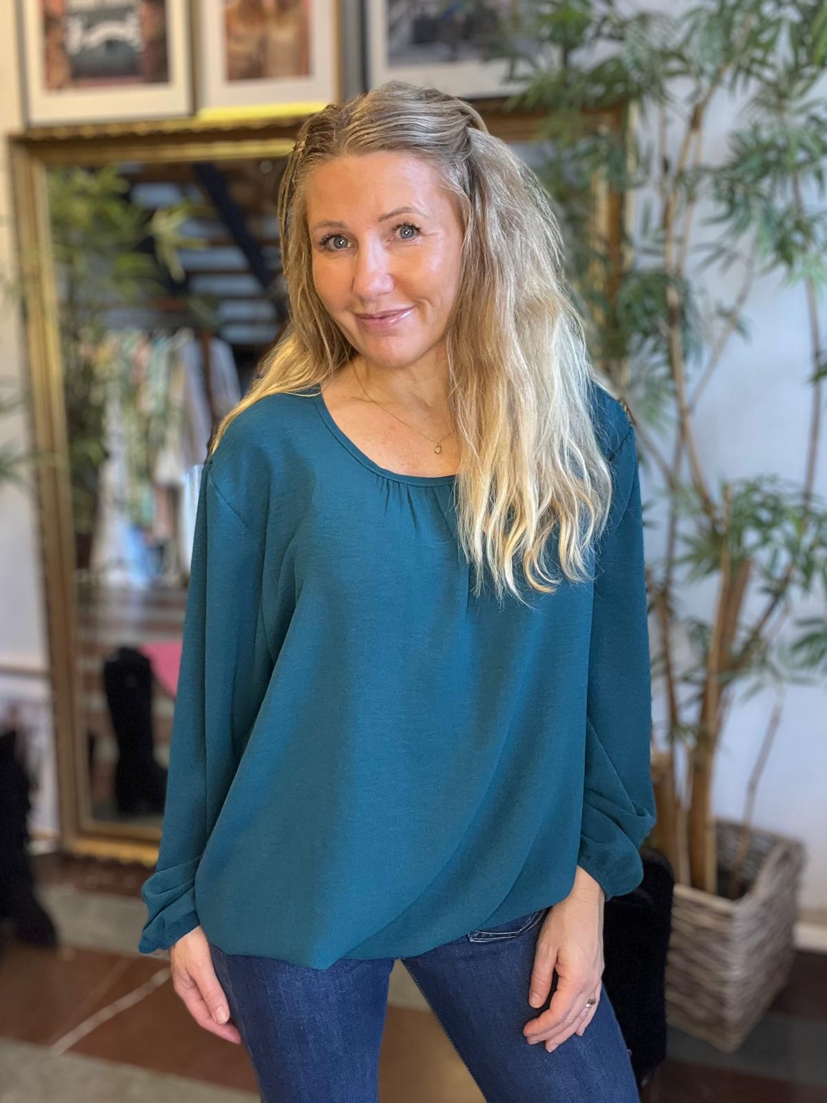 Kvinna med långt blont hår står och ler mot kameran, klädd i en petrolfärgad blus med långa ärmar och blå jeans. Hon poserar inomhus framför en stor spegel, växter och ramar på väggen.