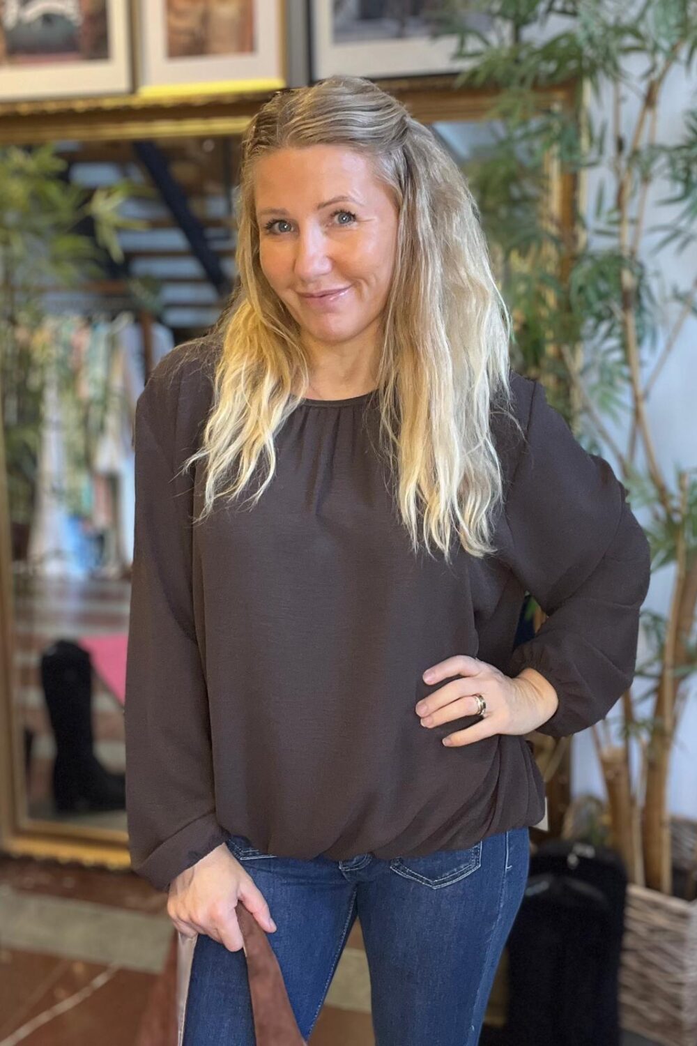 Leende kvinna med långt blont hår står och poserar med ena handen på höften och den andra på en stolrygg. Hon bär mörk blus och jeans, i ett inomhusrum med speglar, tavlor och en stor växt.