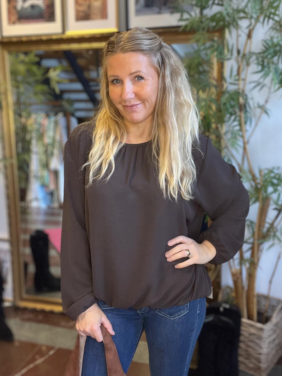 Leende kvinna med långt blont hår står och poserar med ena handen på höften och den andra på en stolrygg. Hon bär mörk blus och jeans, i ett inomhusrum med speglar, tavlor och en stor växt.