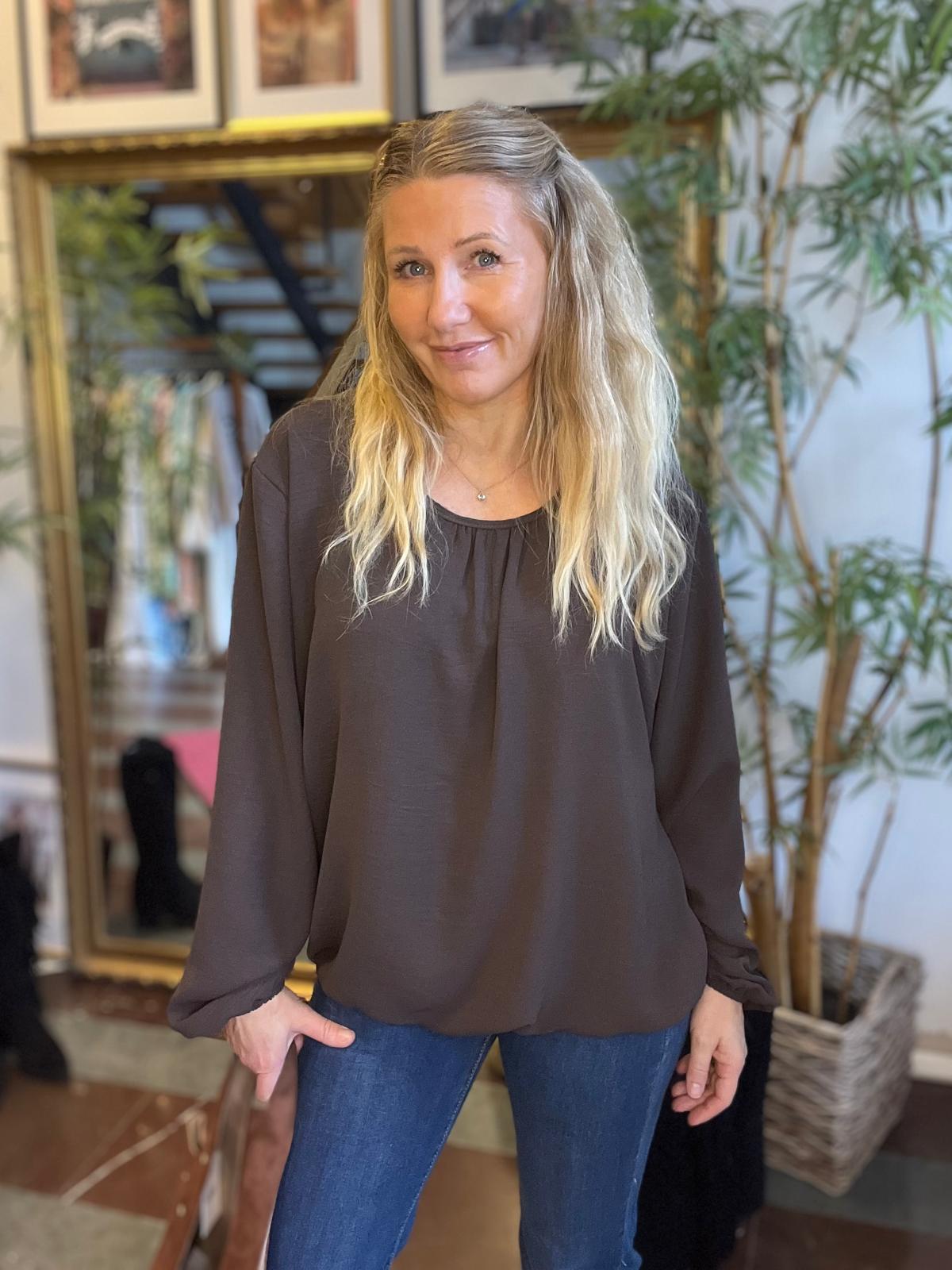 Blond kvinna står och ler mot kameran med ena handen vid höften, klädd i mörkblus och blå jeans. Hon befinner sig inomhus framför en stor spegel, inramade tavlor och en hög grön växt.
