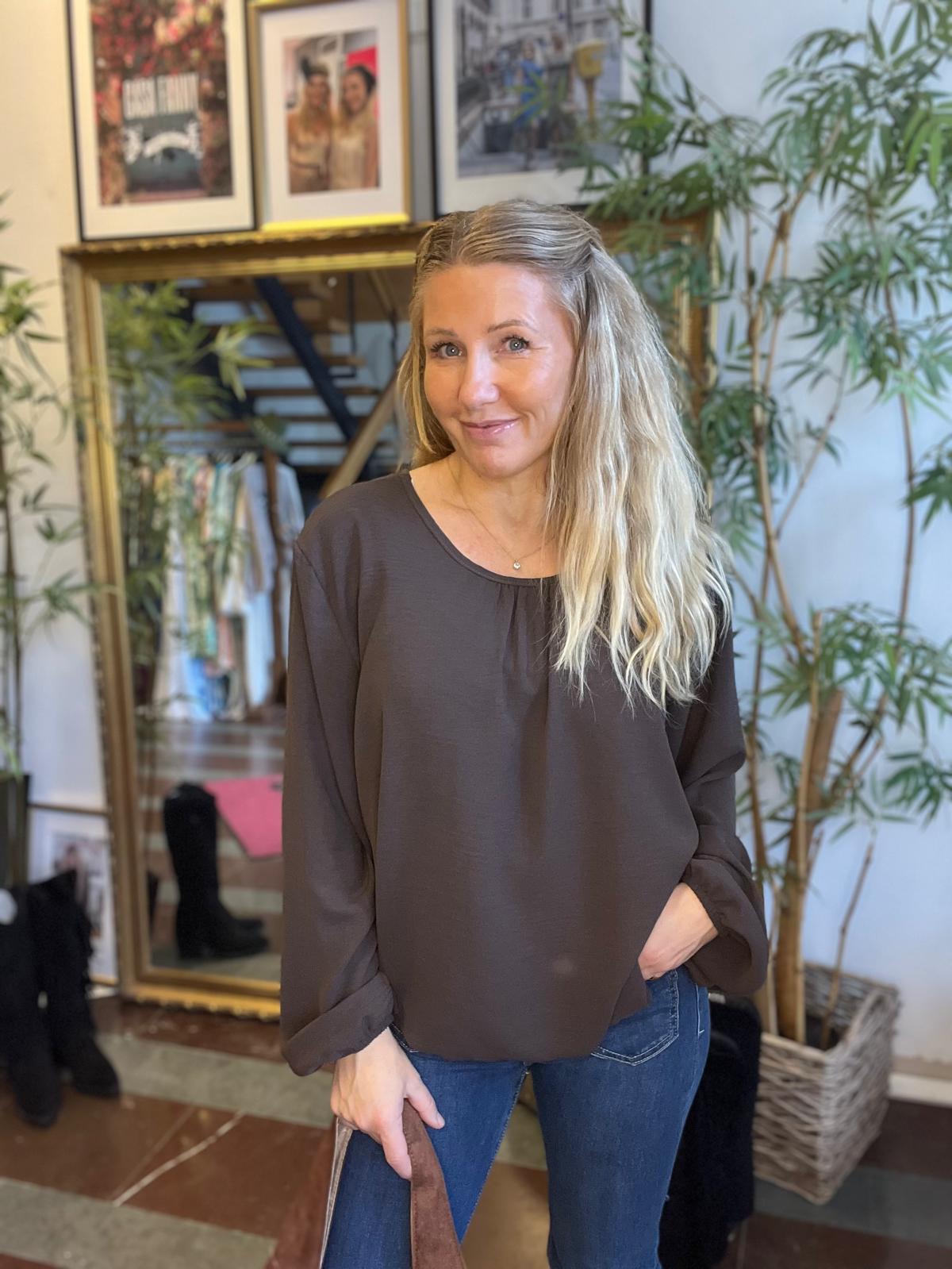 Kvinna med långt blont hår ler mot kameran och står avslappnat med en hand i jeansfickan, klädd i mörk blus och blå jeans. Inomhusmiljö med stor spegel, inramade foton, kläder och växter i bakgrunden.