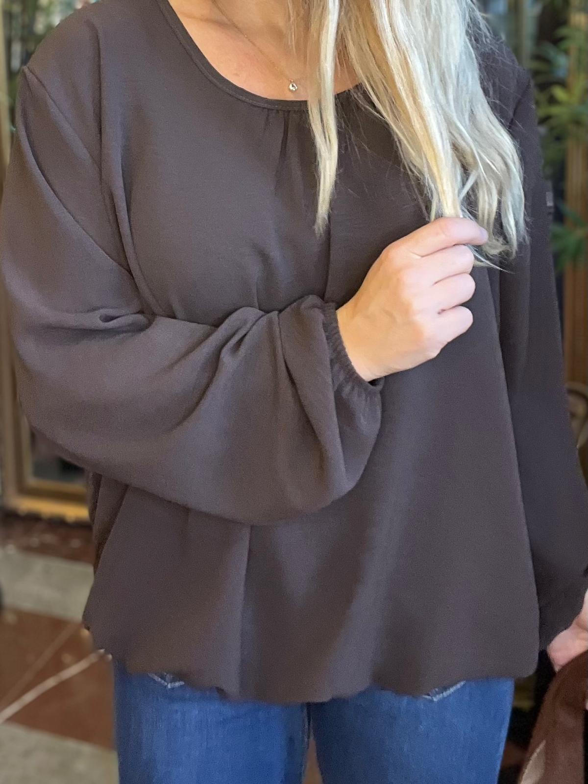Kvinna i mörkgrå, lös blus med vida ärmar och rund hals ringad, håller en slinga av sitt blonda hår i handen. Närbild inomhus, med jeans synliga och en liten halskedja.