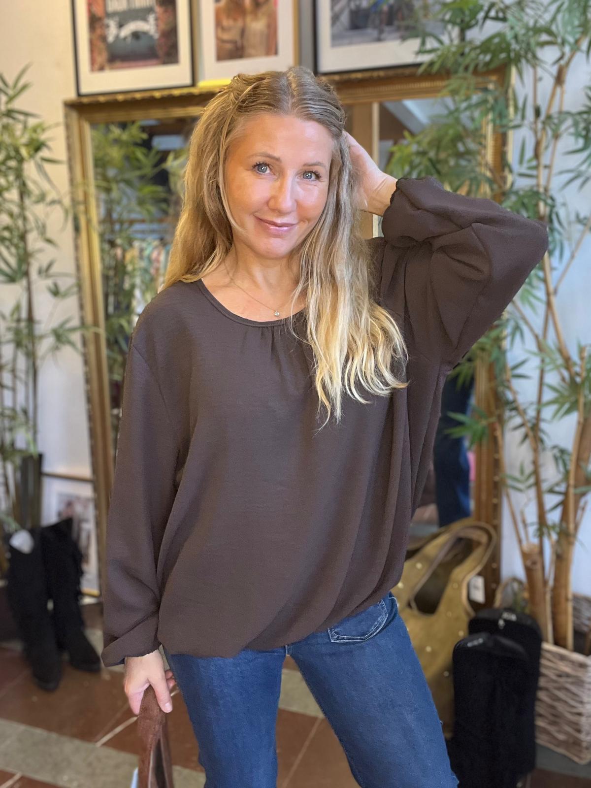 Leende kvinna med långt blont hår poserar med ena handen bakom huvudet och den andra vid en brun läderväska. Hon bär mörk blus och jeans, i ett rum med spegel, växter och inramade foton.
