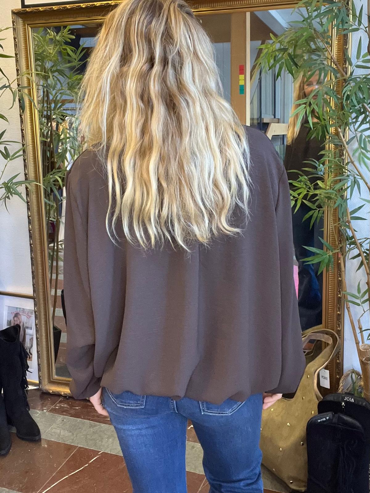 Person med långt, vågigt blont hår står med ryggen mot kameran och tittar i en stor guldramsspegel. Hen bär mörk blus och blå jeans, omgiven av gröna krukväxter i en butik eller salong.