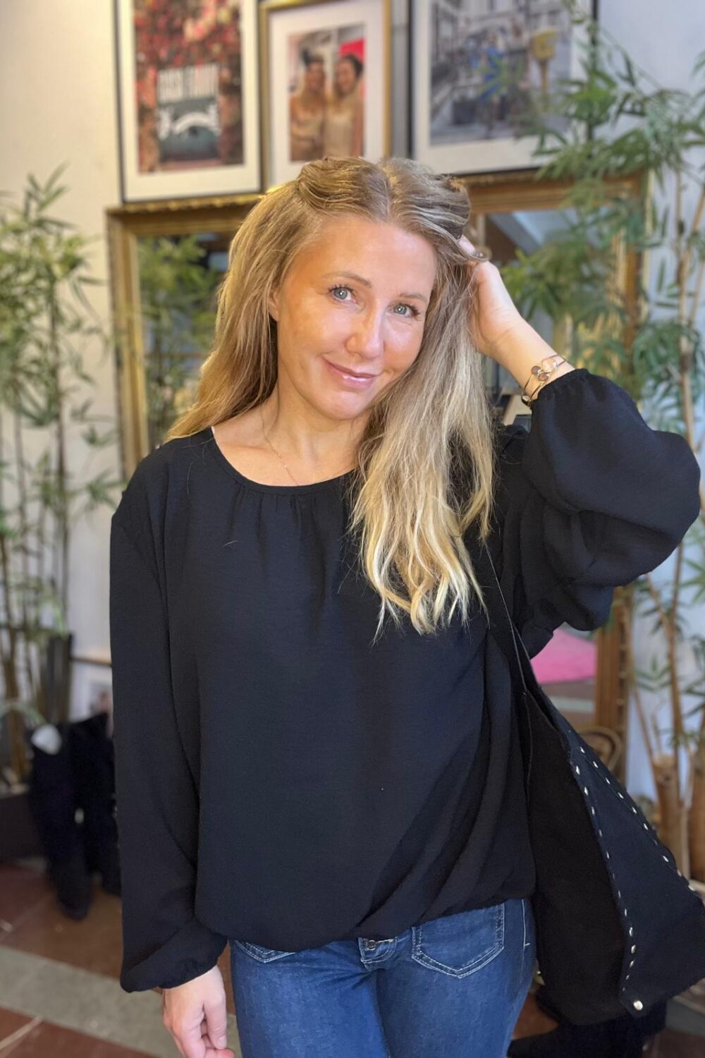 Leende kvinna med långt blont hår står inomhus och håller ena handen i håret. Hon bär svart blus och jeans samt en mörk jacka över axeln, framför växter och inramade tavlor på väggen.