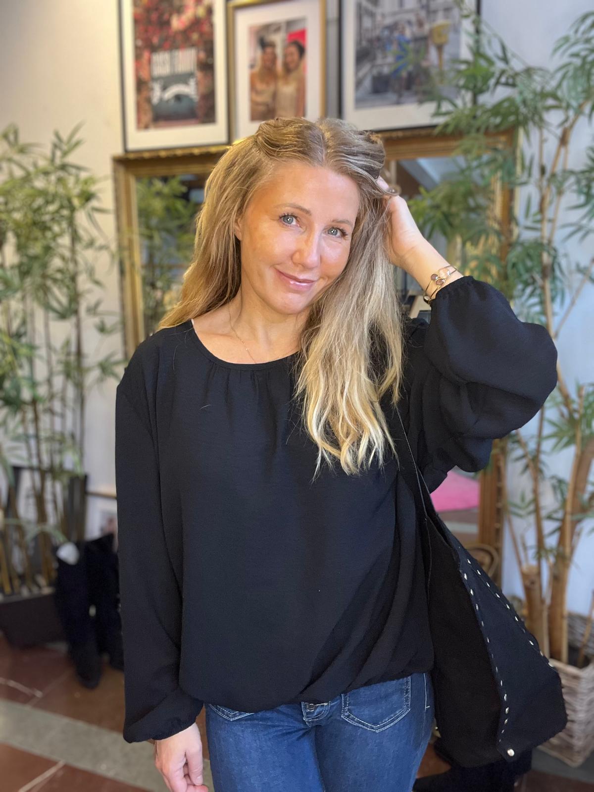 Leende kvinna med långt blont hår står inomhus och håller ena handen i håret. Hon bär svart blus och jeans samt en mörk jacka över axeln, framför växter och inramade tavlor på väggen.
