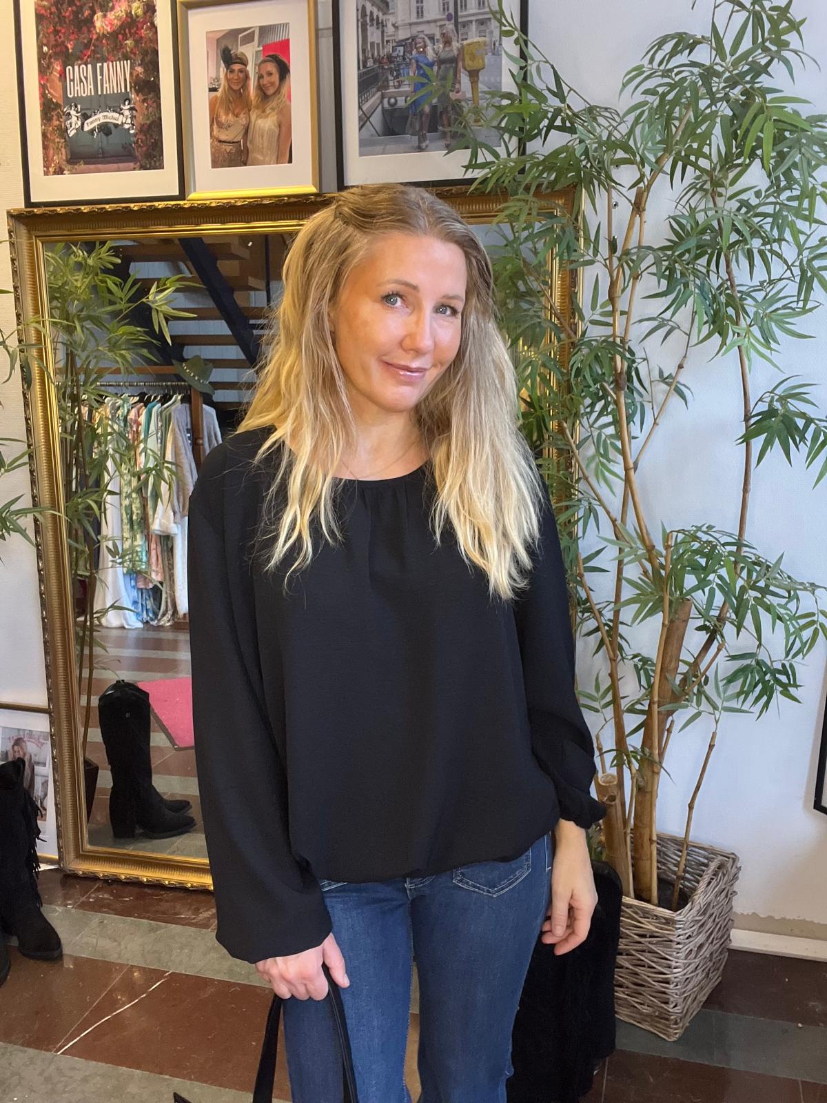 Blond kvinna står och ler lätt mot kameran i en butiksmiljö, klädd i svart blus och blå jeans. Bakom henne syns en stor guldramsspegel, klädhängare, bambuväxt och inramade foton med texten “CASA FANNY”.