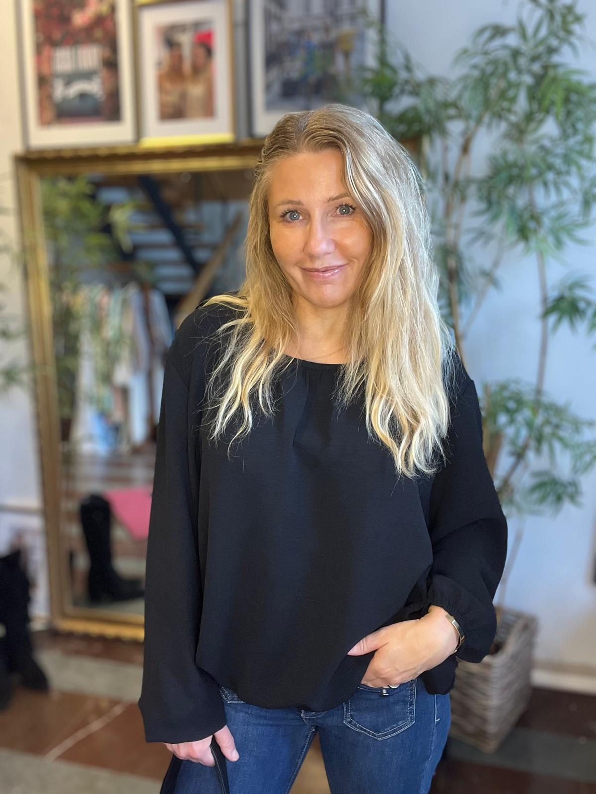 Blond kvinna i svart långärmad blus och jeans står och ler mot kameran, med ena handen i fickan. Inomhusmiljö med stor spegel, inramade tavlor och grön växt i bakgrunden.