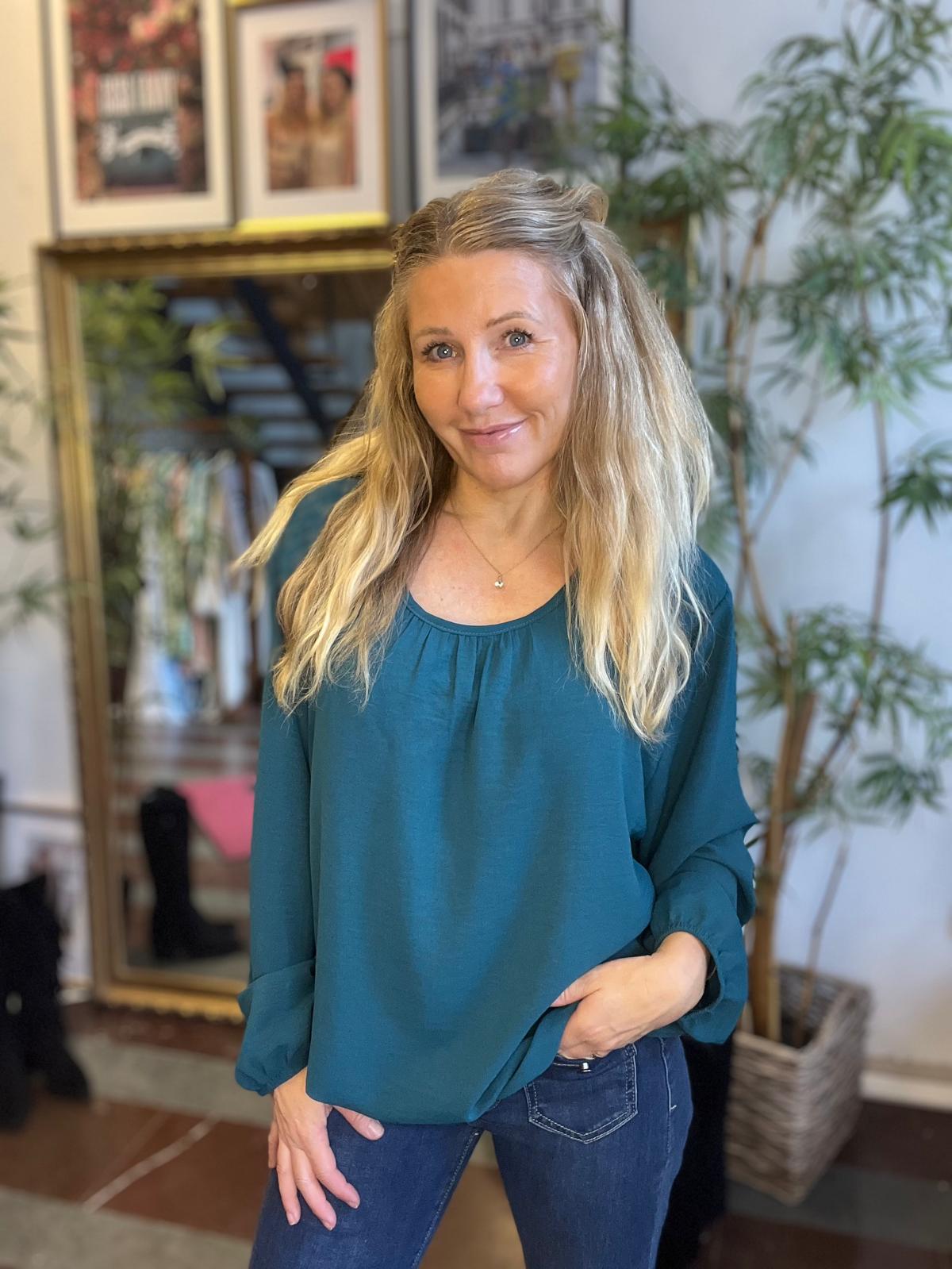 Leende kvinna med långt blont hår står och poserar inomhus, med ena handen i jeansfickan och den andra vid tröjans kant. Hon bär en grönblå blus; bakom syns en stor spegel, inramade foton och växter.