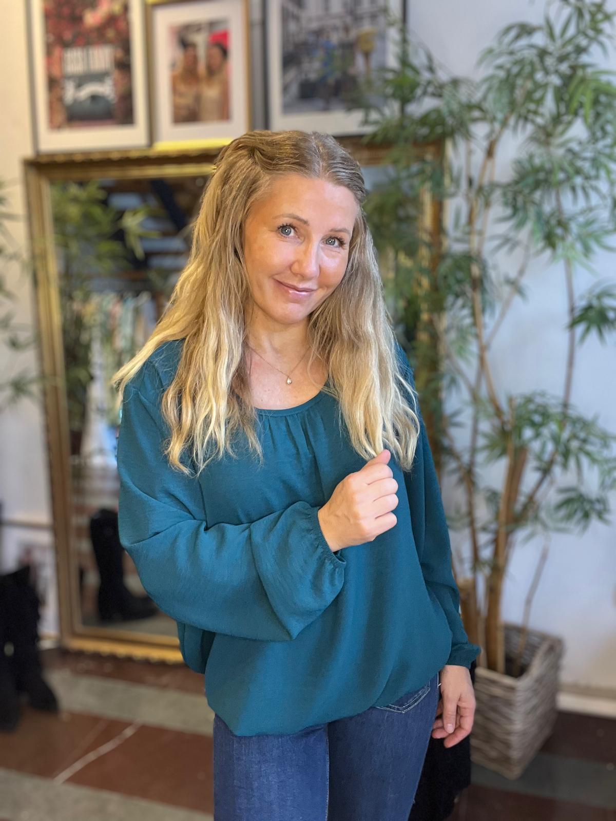 Kvinna med långt blont hår står och ler mot kameran med ena handen lätt upplyft vid bröstet. Hon bär turkos blus och jeans, inomhus framför en guldramsspegel, inramade foton och en stor grön växt.