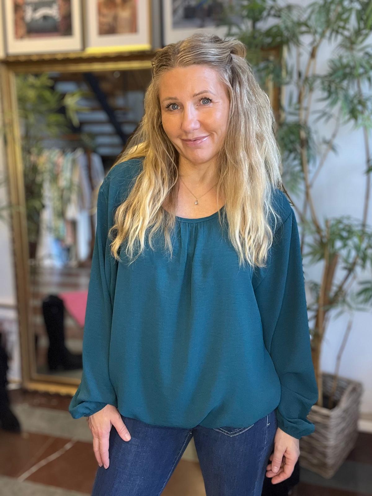 Leende kvinna med långt blont hår står och lutar sig lätt framåt, med händerna vid låren, iklädd en turkos blus och jeans. Inomhusmiljö med spegel, trappa och gröna växter i bakgrunden.