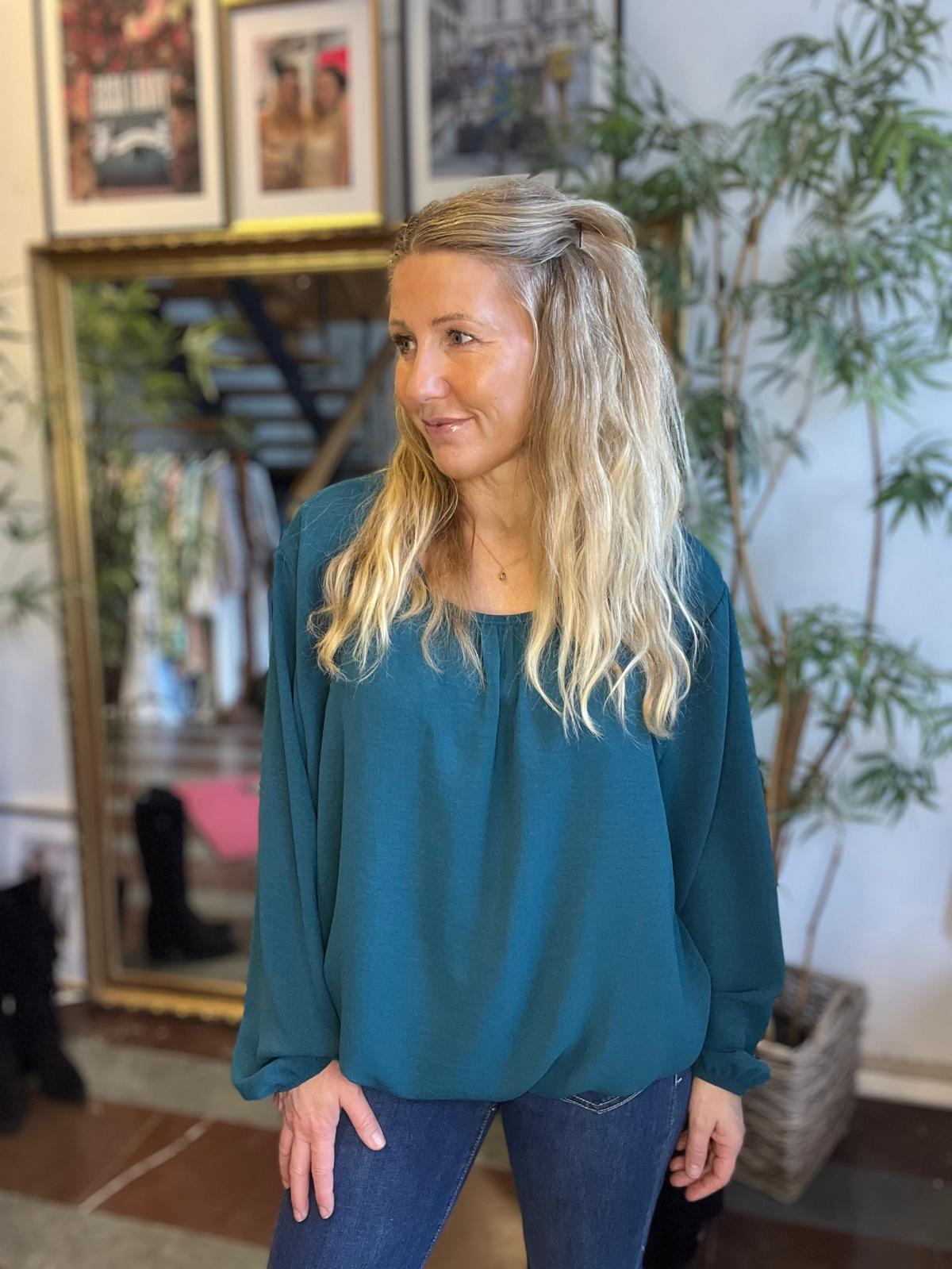 Blond kvinna står inomhus och tittar åt sidan med ett svagt leende, iklädd blågrön blus och jeans. I bakgrunden syns spegel, inramade foton på väggen och en stor grön växt.