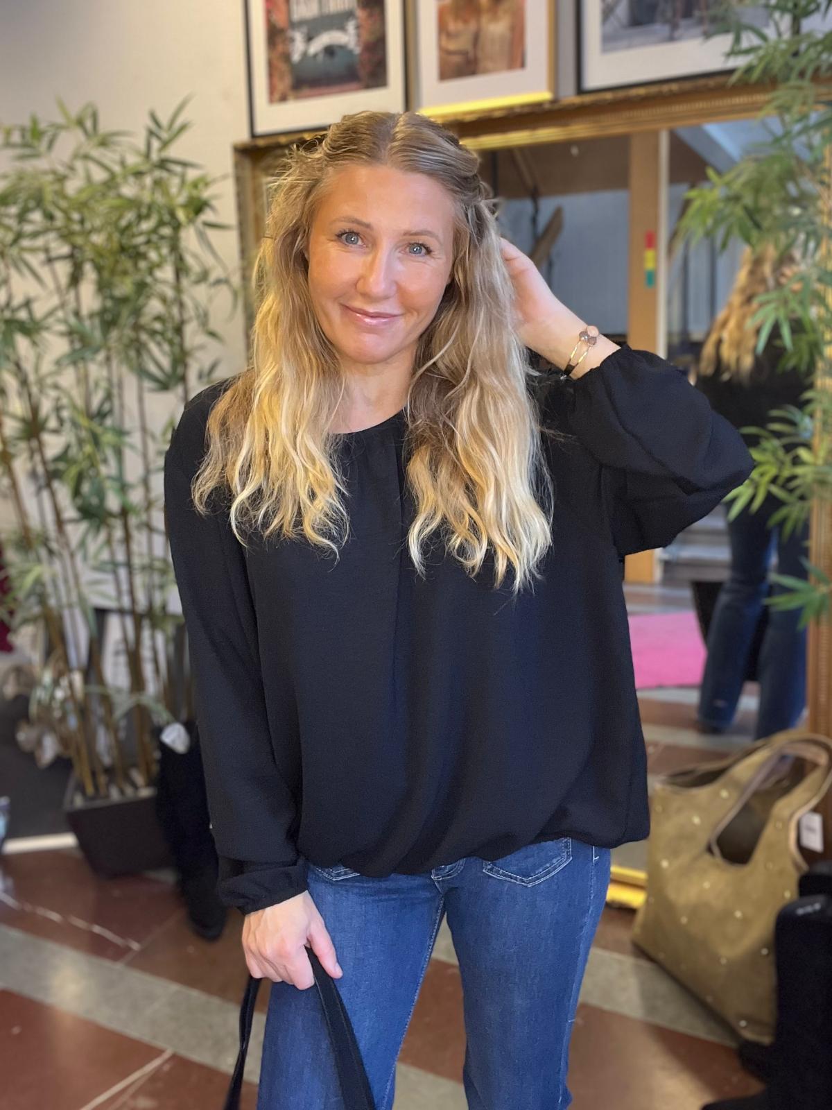 Leende kvinna med långt blont hår står inomhus och håller en väska i ena handen medan hon rör vid håret med den andra. I bakgrunden syns gröna växter, en stor spegel och inramade tavlor.