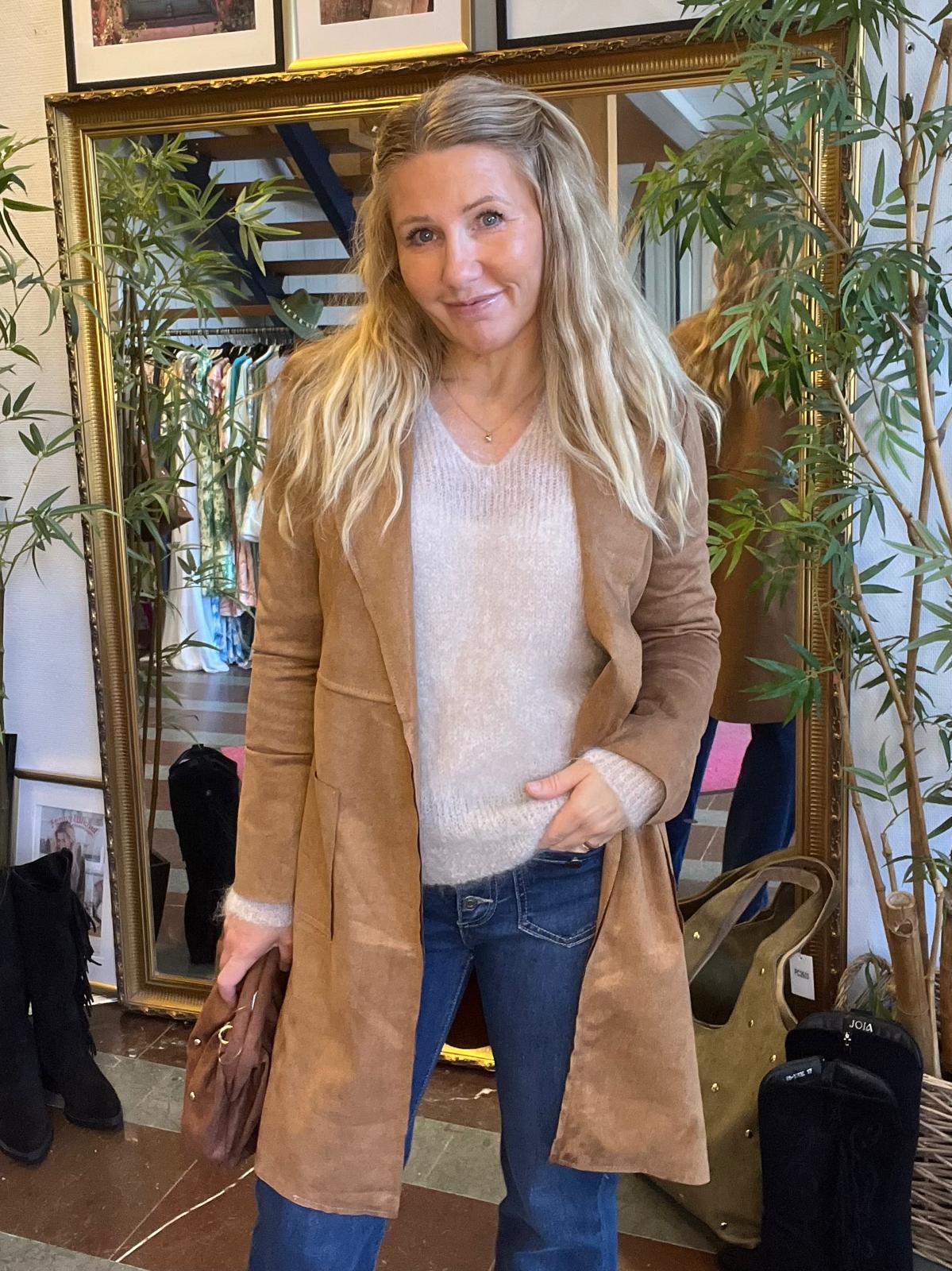 Kvinna med långt blont hår står och ler framför en stor guldramsspegel, klädd i beige kappa, ljus stickad tröja och jeans, med en liten brun väska i handen. Spegeln visar klädställning och grön växt.