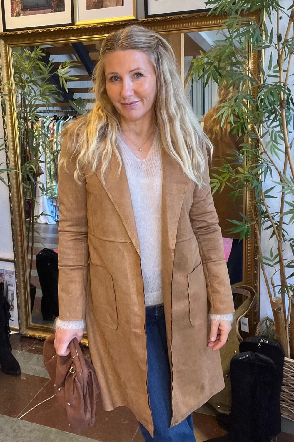 Kvinna med långt blont hår står och ler mot kameran, klädd i beige mockakappa över ljus tröja och jeans, och håller en brun väska. Hon står framför en stor spegel och växter i en butiksmiljö.