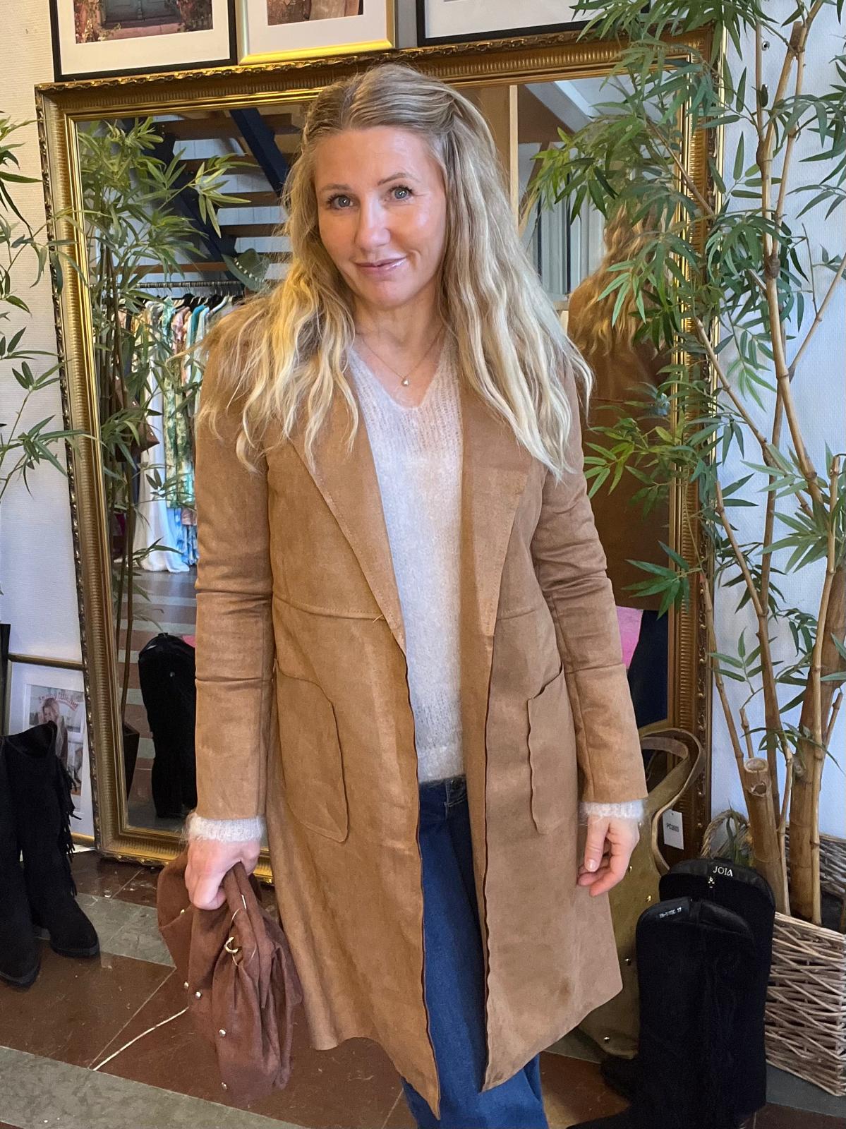 Kvinna med långt blont hår står och ler mot kameran, klädd i beige mockakappa över ljus tröja och jeans, och håller en brun väska. Hon står framför en stor spegel och växter i en butiksmiljö.