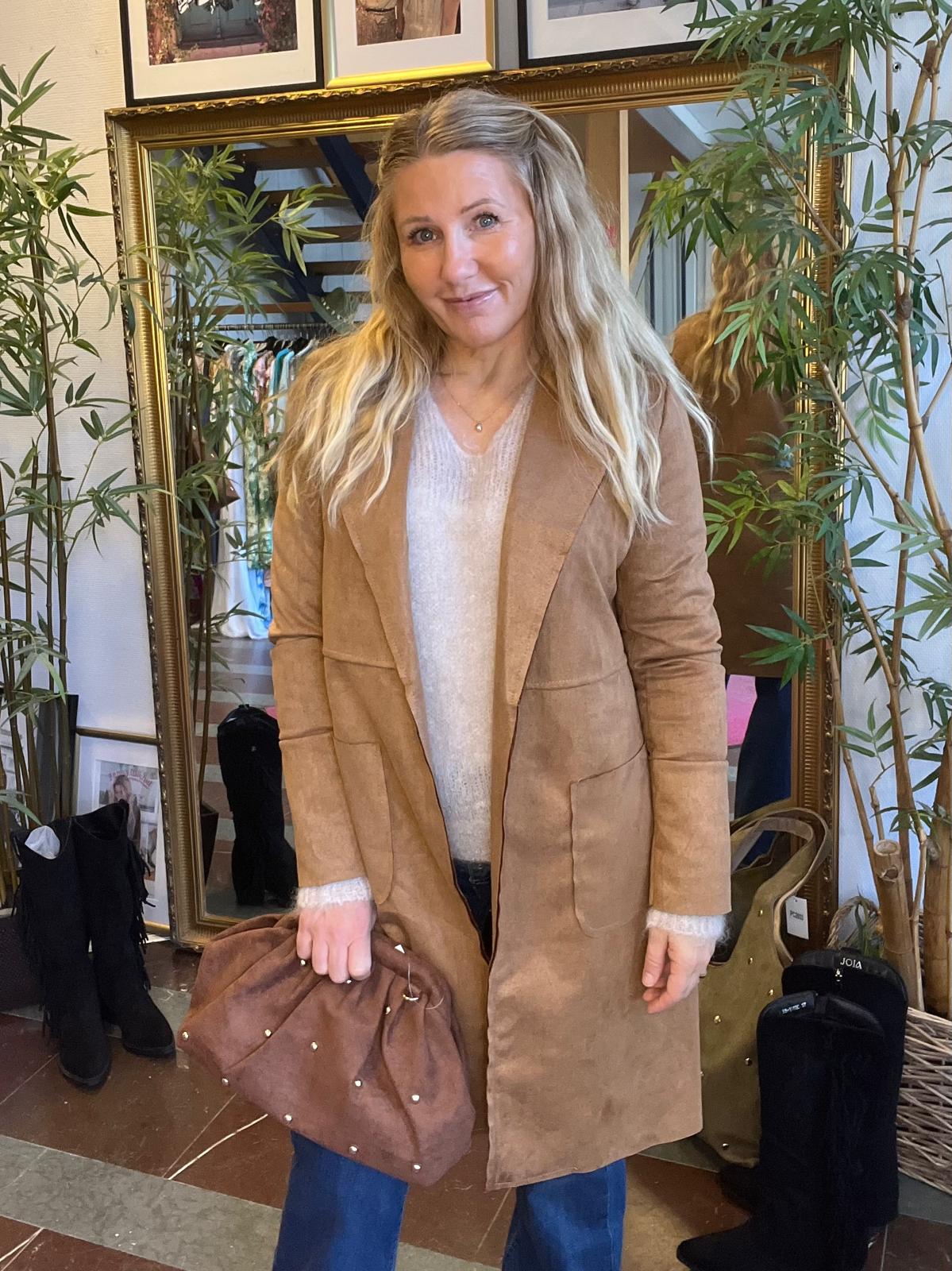 Leende kvinna står och poserar framför en stor guldramsspegel, iklädd lång brun mockakappa och ljus stickad tröja, med en brun väska i handen. I bakgrunden syns klädställning, skor och gröna krukväxter.